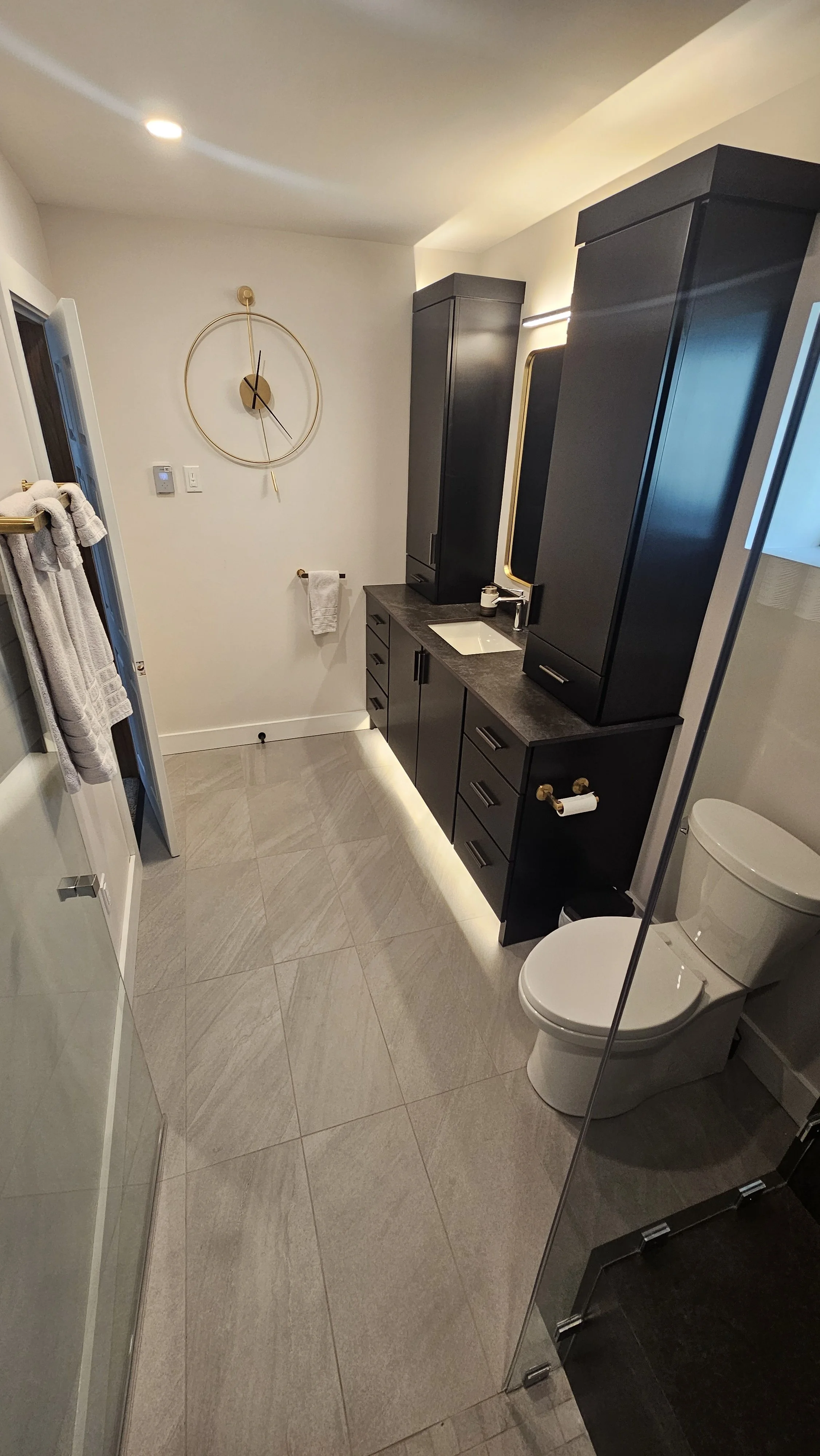 A modern bathroom with black cabinetry, a white toilet, beige tile floor, and a wall clock with gold accents.