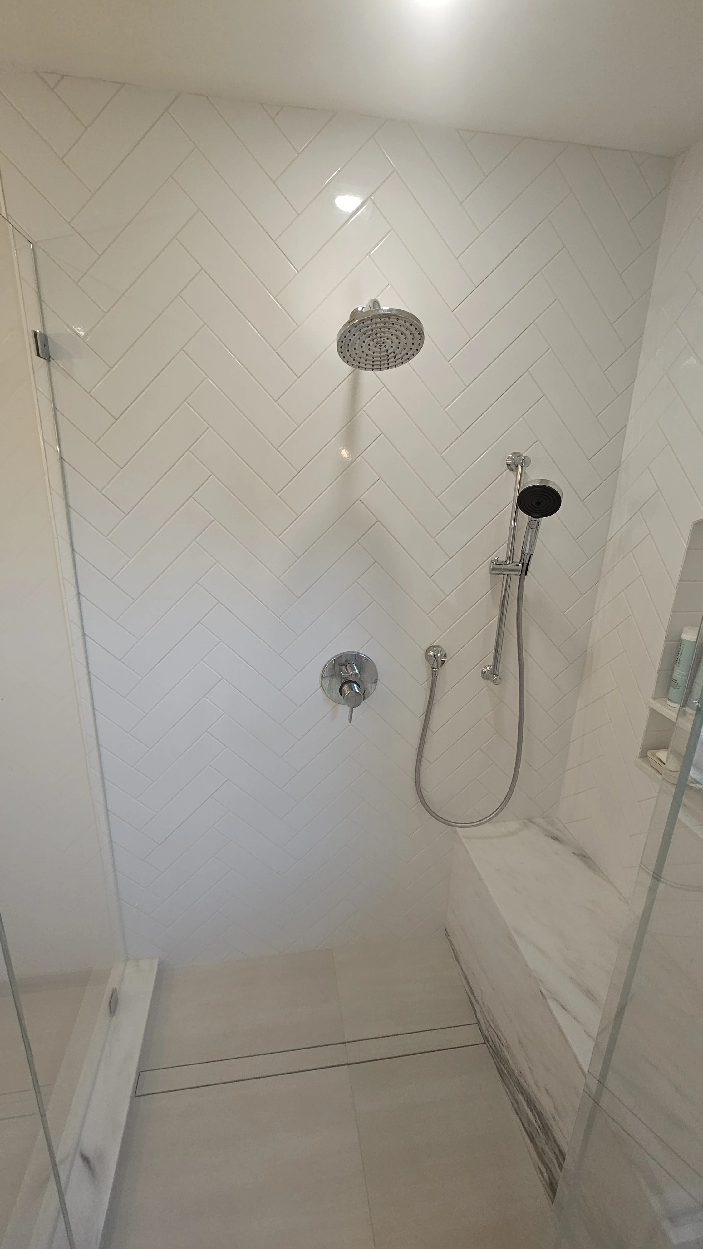 Bathroom shower with white herringbone tile wall, large showerhead, and handheld shower attachment on marble ledge.