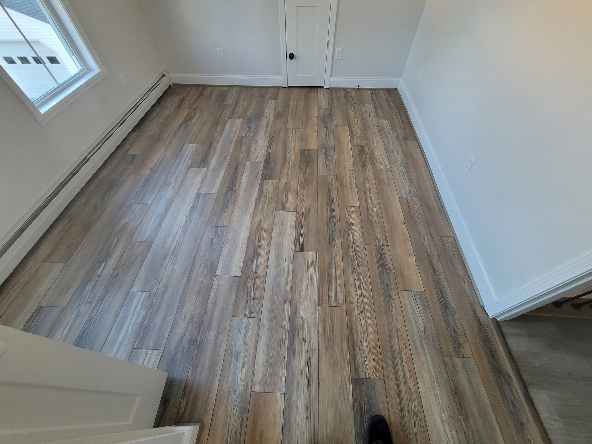 A room with hardwood flooring, a window on the left, white walls, baseboard heating, and a closed door on the back wall.