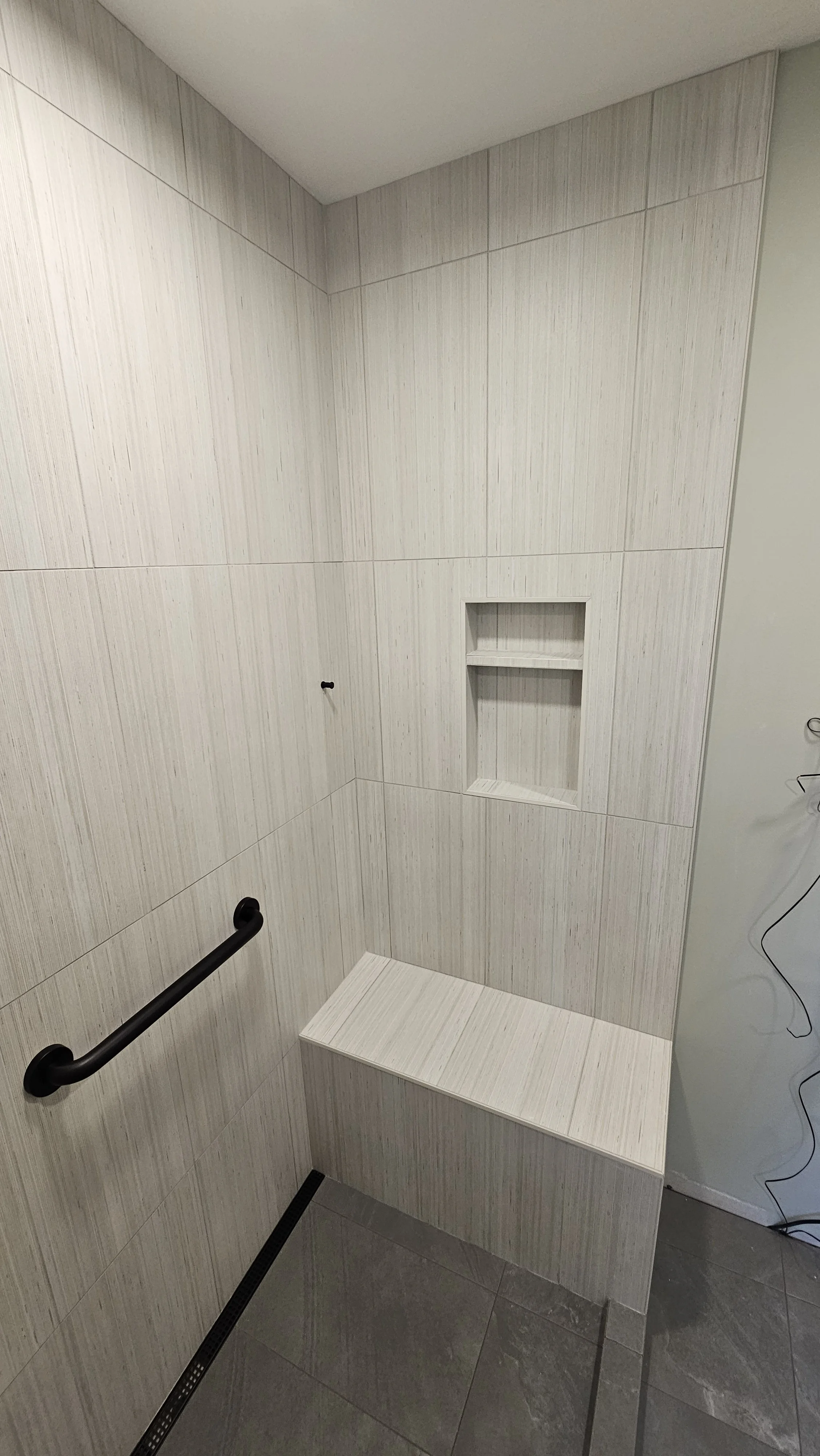 A shower area with beige vertical striped tiles, a black safety grab bar, a built-in niche for toiletries, and a bench at the corner. The floor has darker gray tiles, and there is a linear drain on the floor.