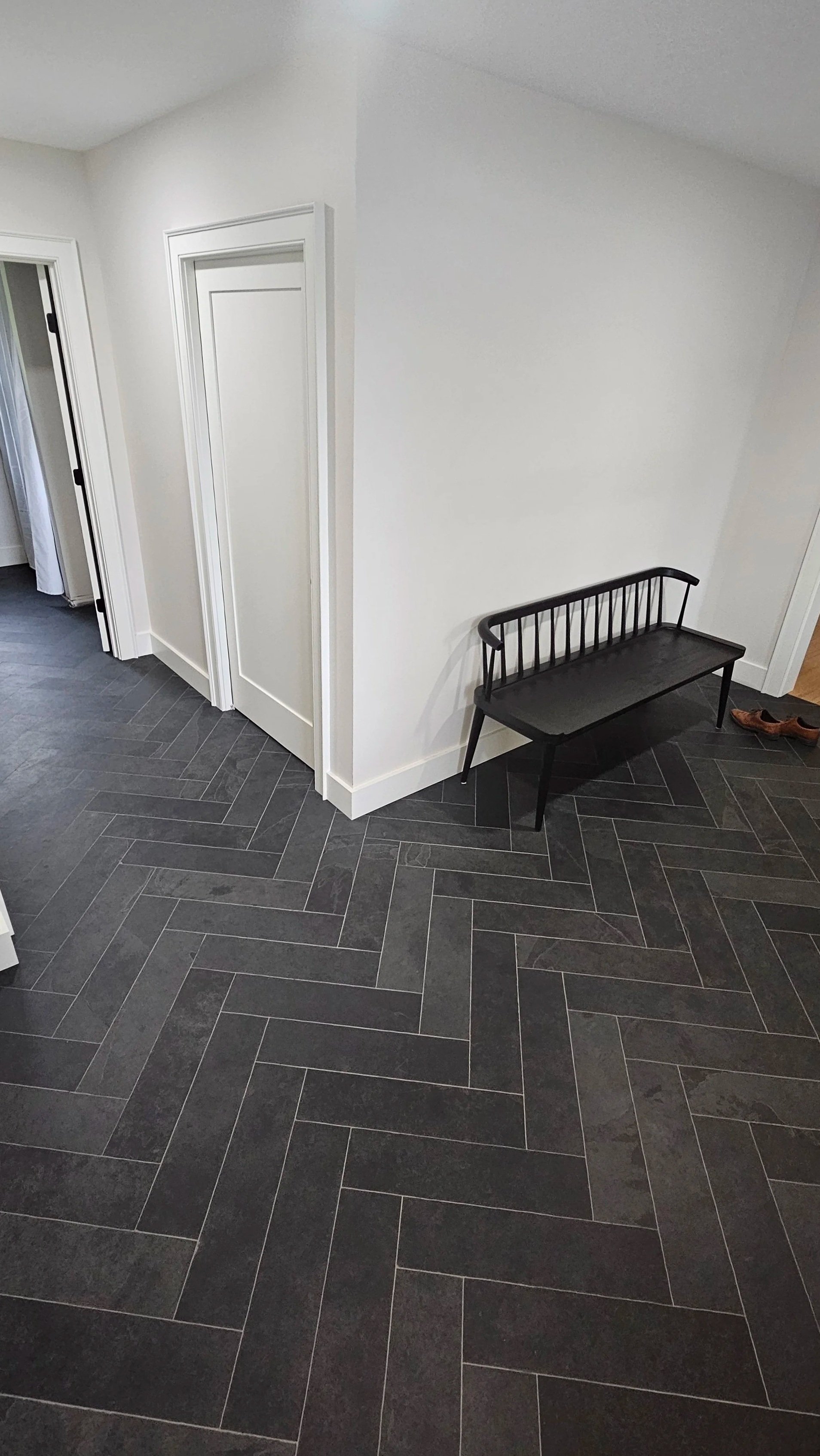 Interior view of a hallway with dark herringbone-patterned tile flooring, white walls, a white door, and a black bench against the wall. Shoes are near the door.