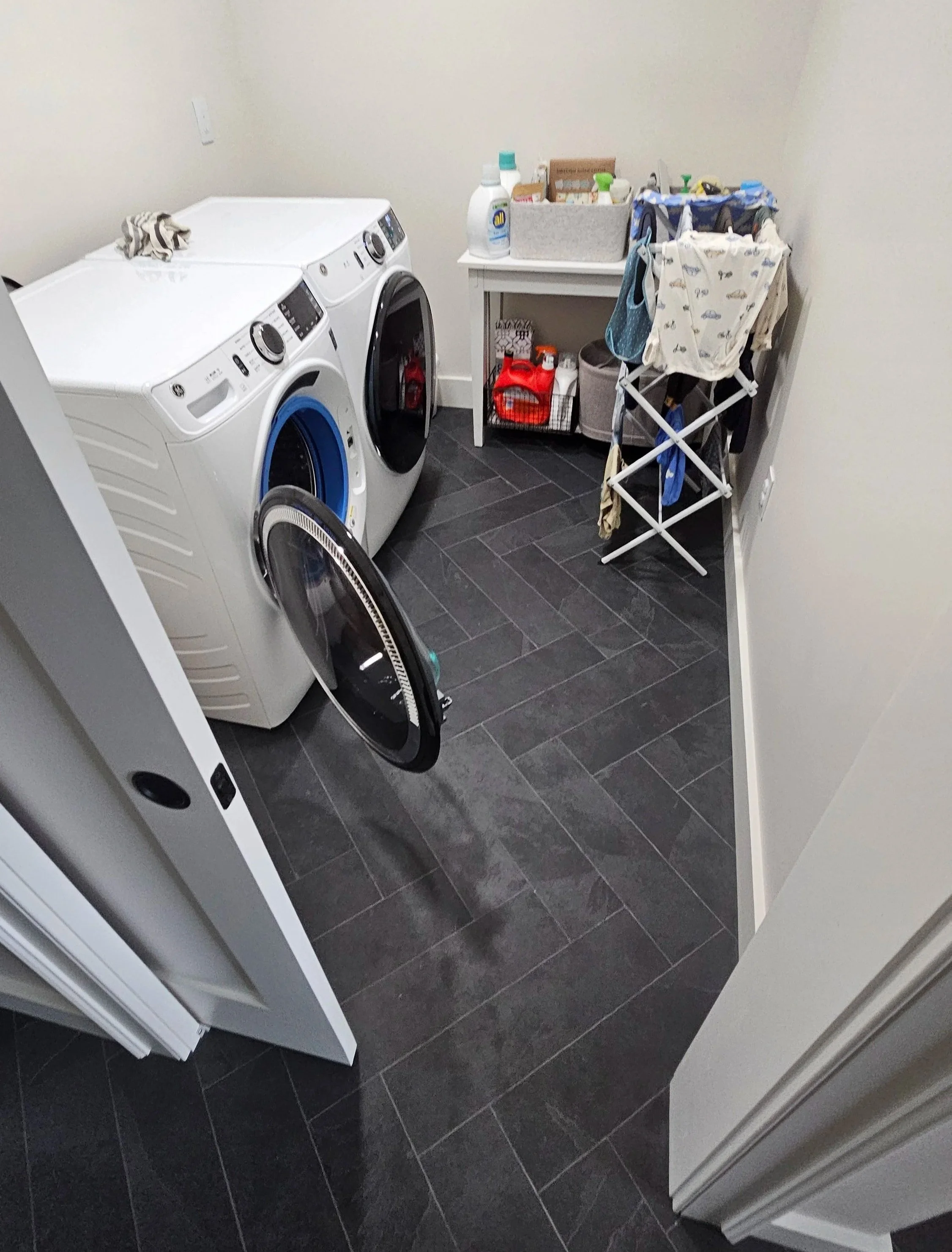 A laundry room with a front-loading washing machine and dryer, a small table with laundry supplies, and a drying rack with clothes hanging on it. The room has dark tiled flooring and plain walls.