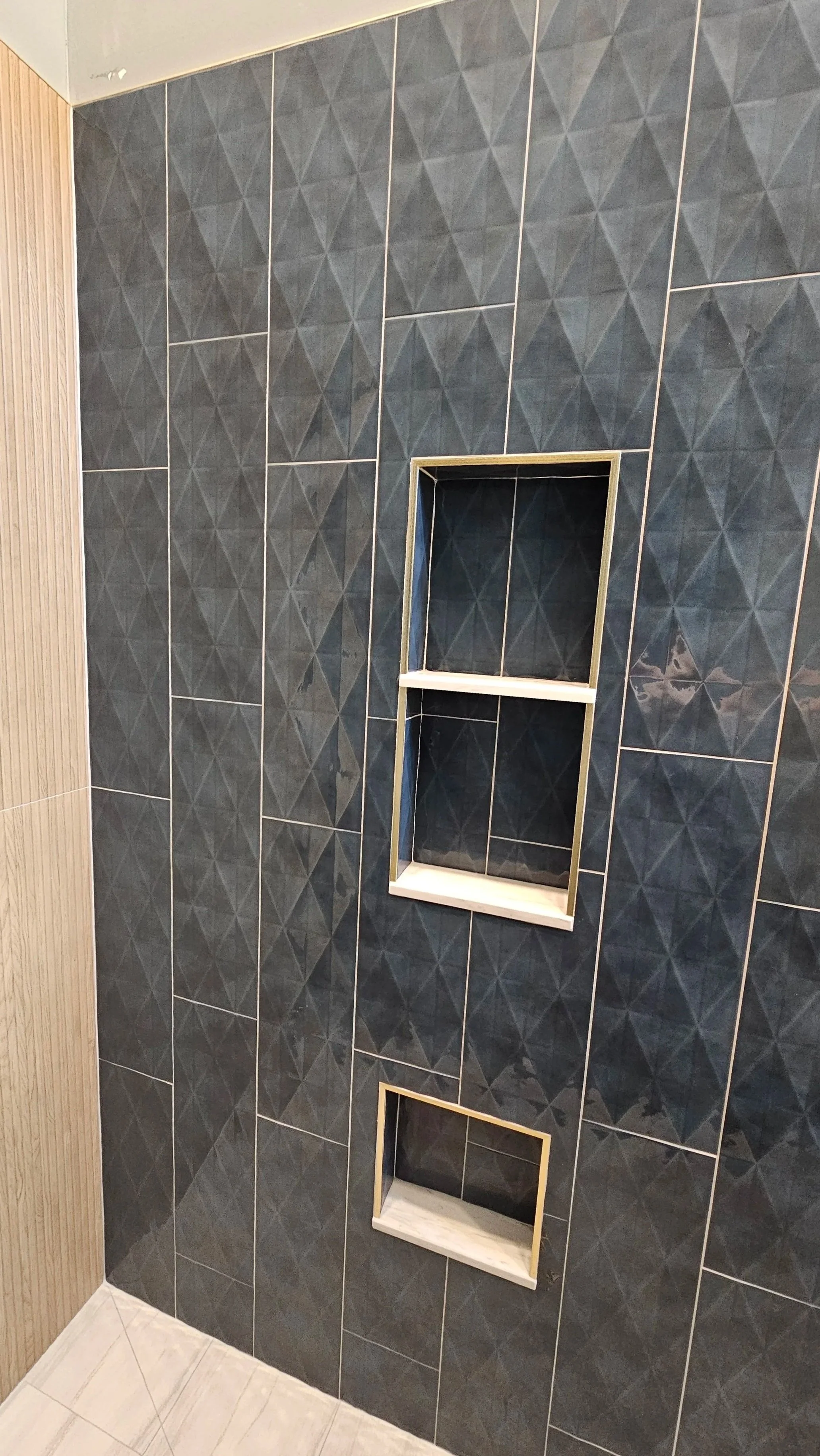 A bathroom shower area with dark gray geometric patterned tiles. There are two built-in shelves framed with light wood, one above the other, in the shower wall. The floor has light-colored tiles.