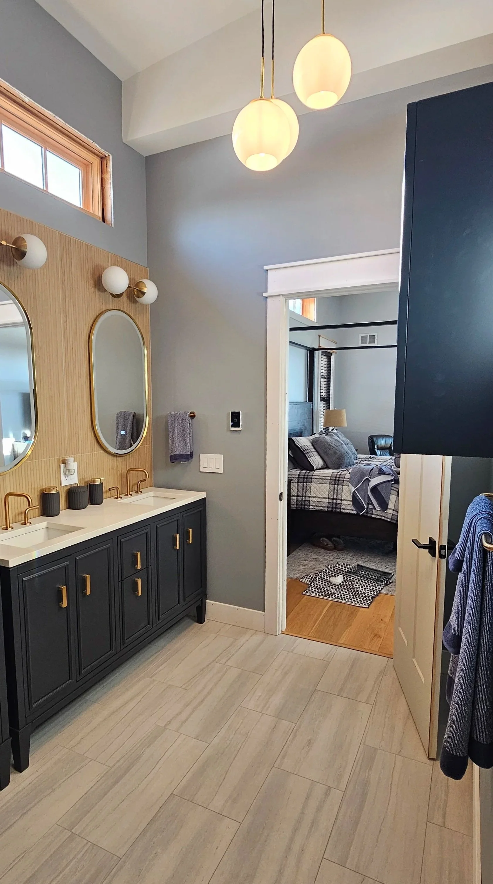 A modern bathroom with a double sink vanity, black cabinets, gold hardware, and oval mirrors. There are three globe light fixtures above the mirrors. A small window is near the ceiling, and a towel rack with a gray towel. Through a doorway, a bedroom