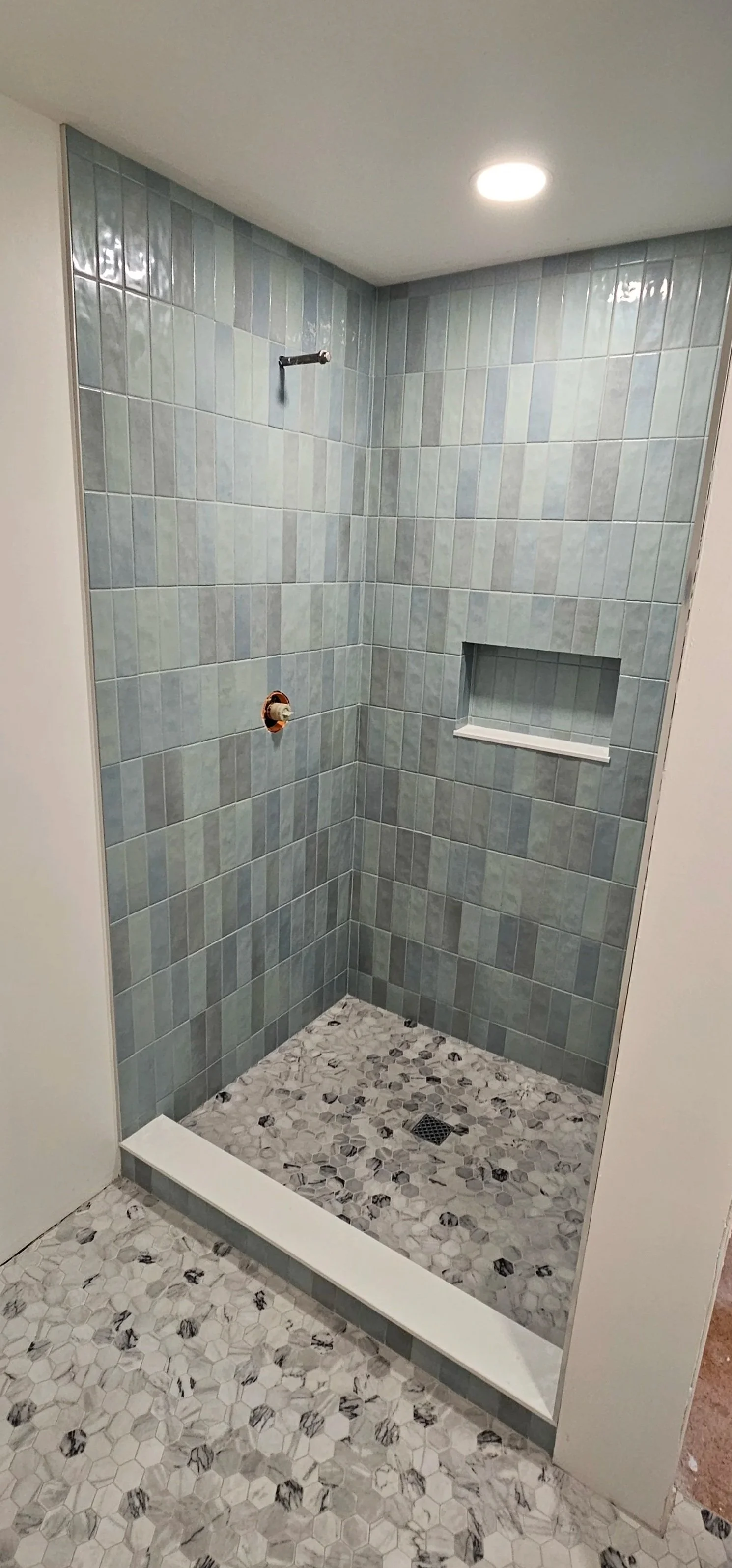 Unfinished walk-in shower with light gray and blue tiles on the walls, a built-in niche, a drain on the floor, and a rough pipe for a showerhead.