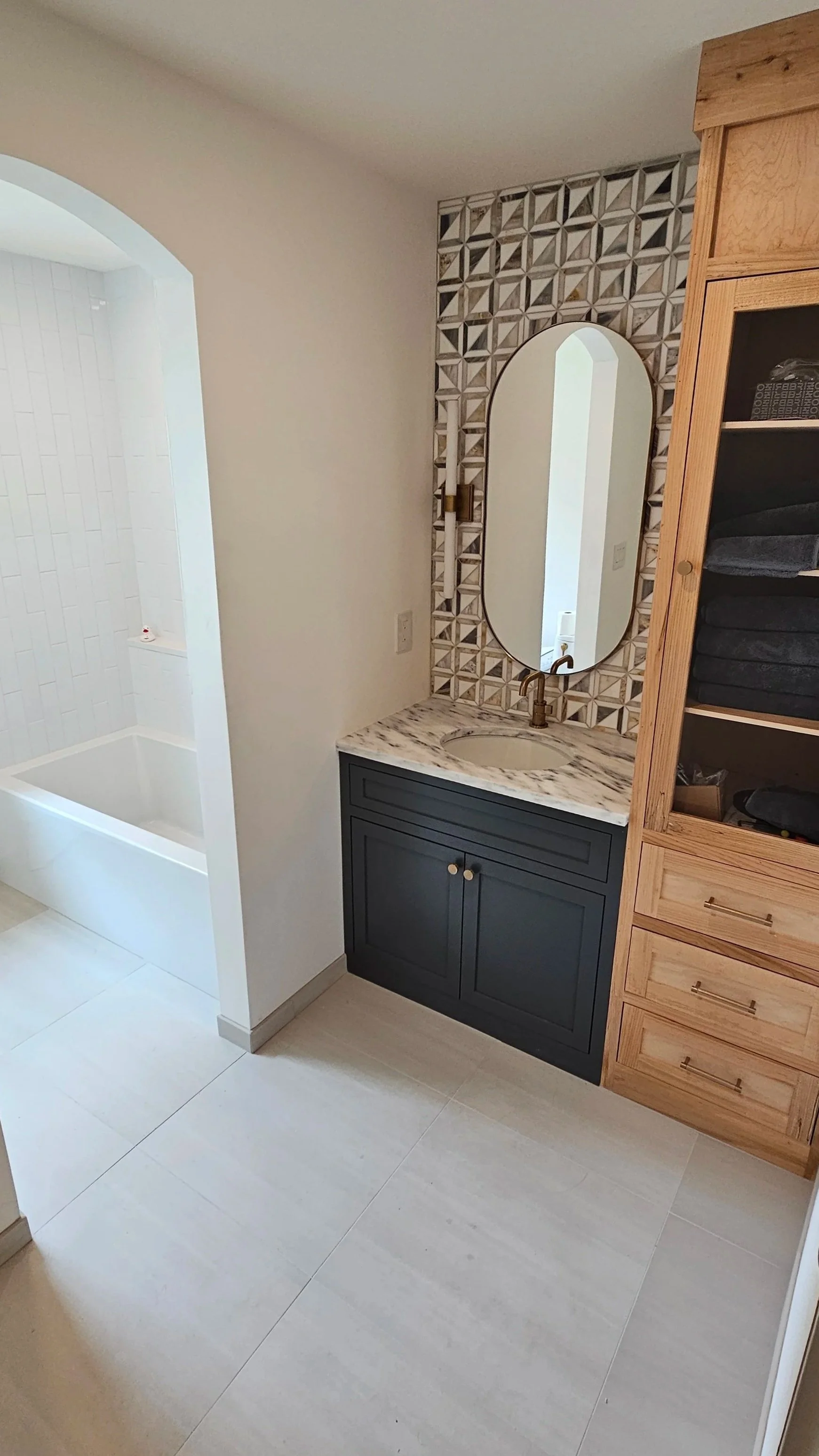 A bathroom with a shower on the left, a dark blue vanity with a marble countertop, and a wooden cabinet with shelves and drawers on the right. There is a decorative tile accent wall behind an oval mirror above the sink.