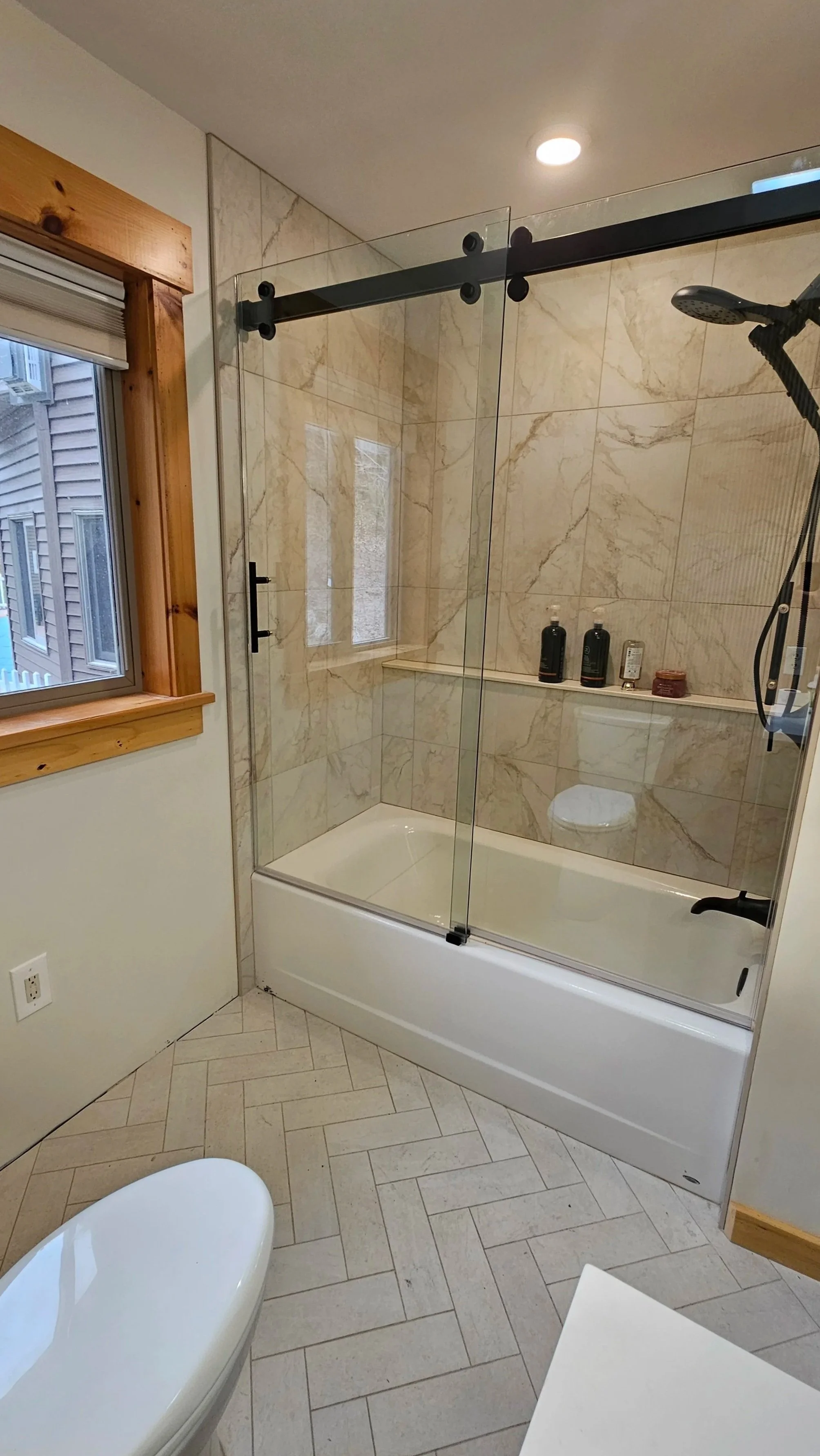 Bathroom with a walk-in shower with beige marble tiles, glass door, wooden window frame, and a white toilet.