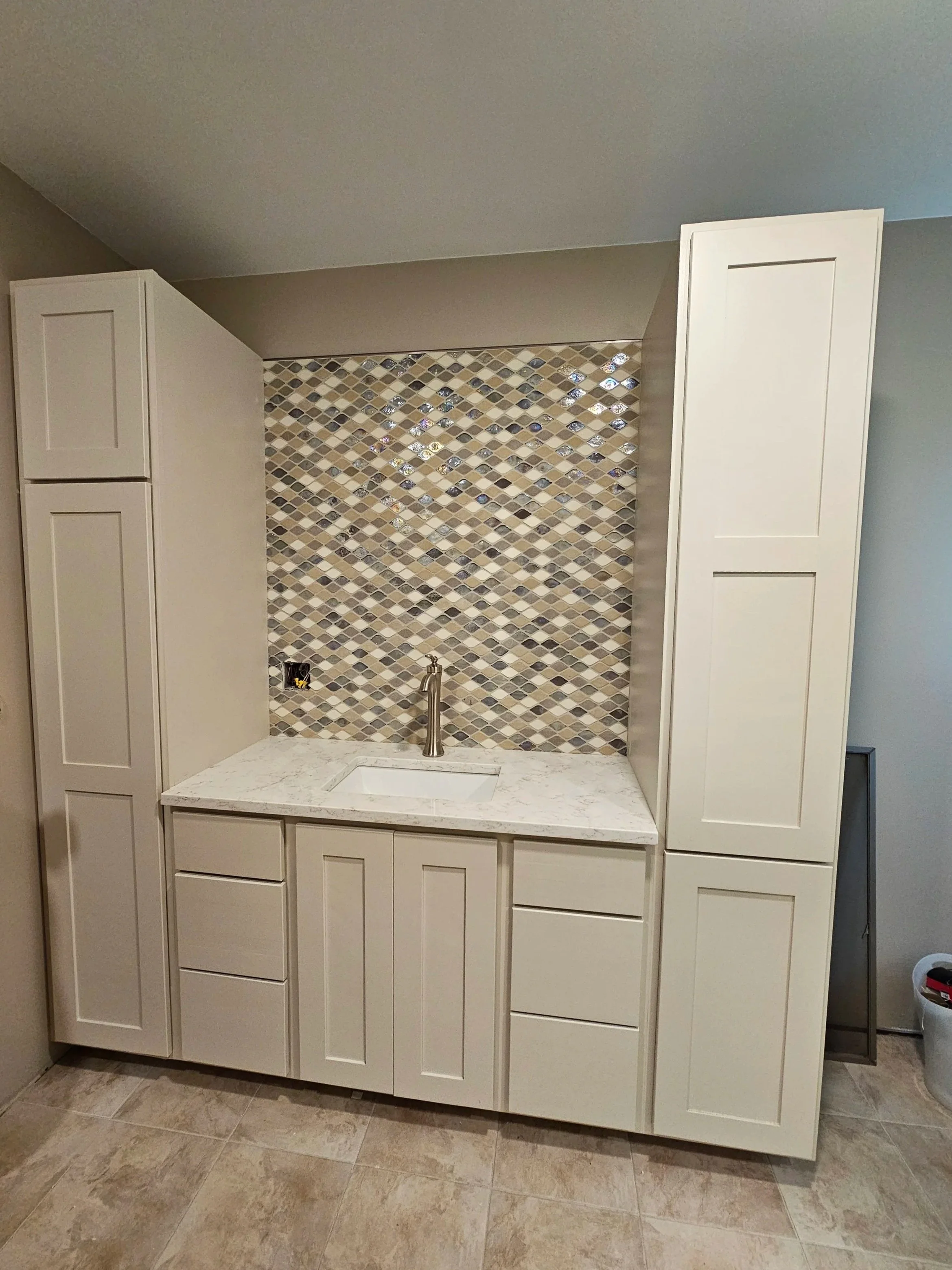 A kitchen with white cabinets surrounding a countertop with a white marble pattern. The backsplash is made of diamond-shaped tiles in shades of beige, brown, and gray with some reflective tiles. There is a small sink with a silver faucet in the cente