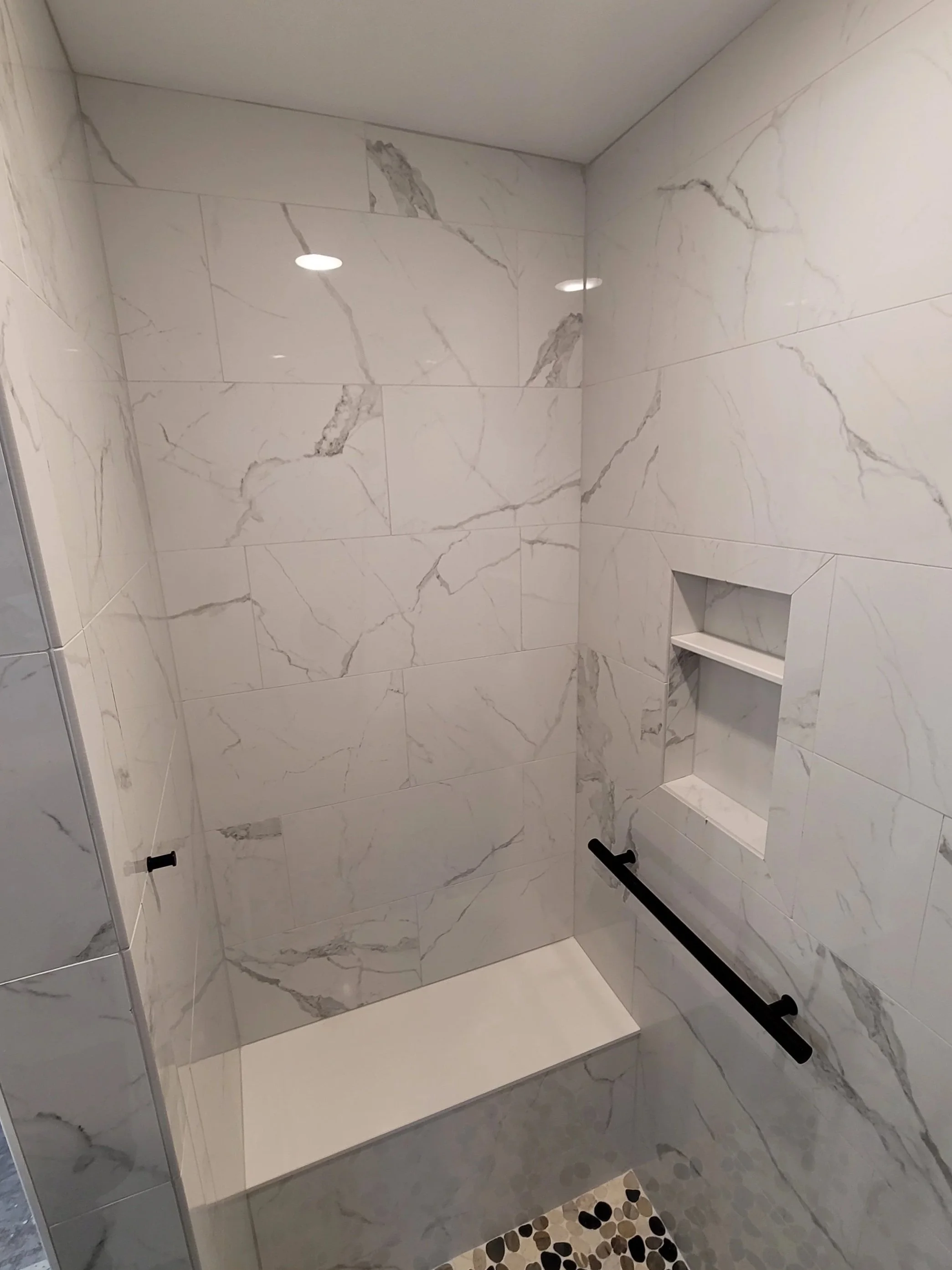 Inside a modern shower with white marble walls, black grab bars, a built-in niche, and a pebble-patterned floor