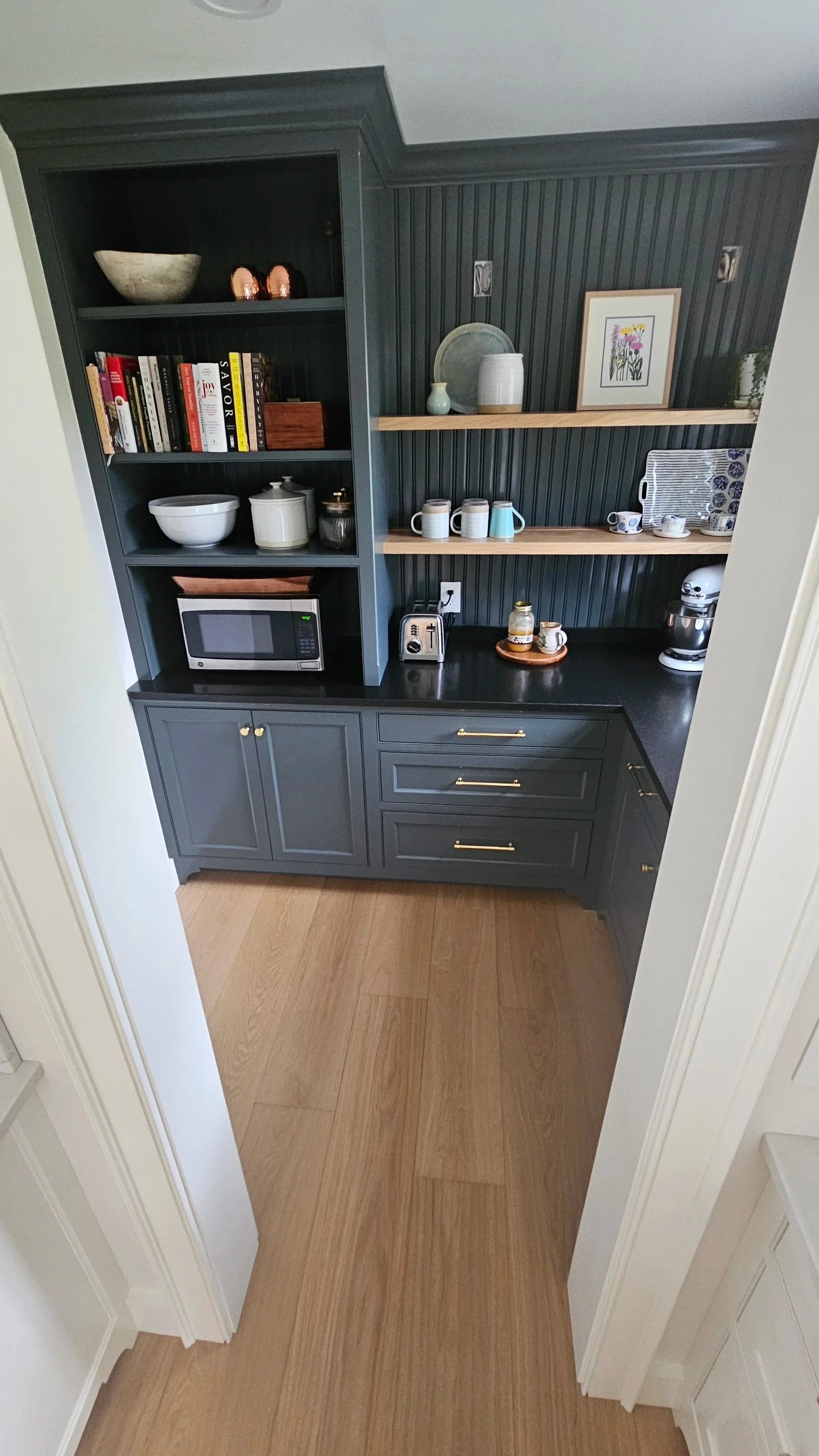 View of a kitchen nook with dark gray built-in cabinetry, open shelving with bowls and books, a microwave, and a countertop with coffee mugs, a toaster, and small jars. Wooden floors and walls with vertical paneling.