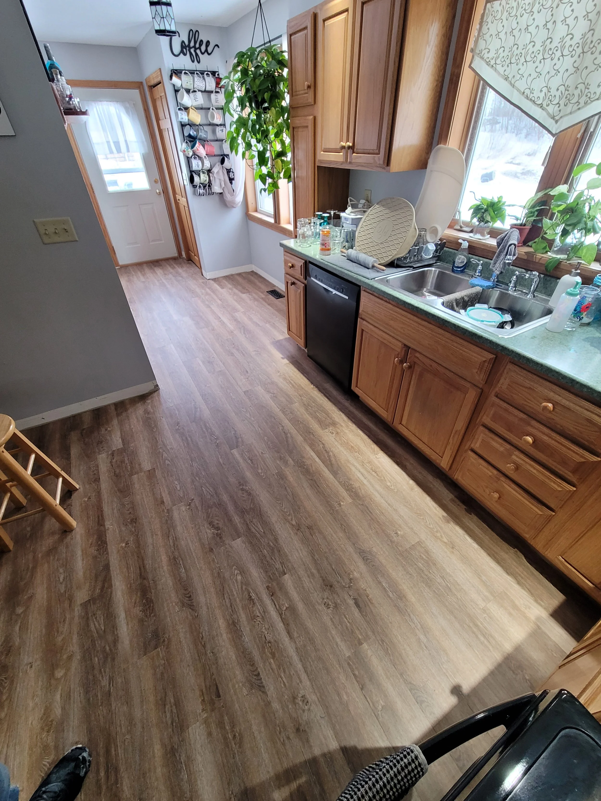 A kitchen with wooden cabinets, a green countertop, a double sink, and large windows with a view of a snowy outdoor scene. There are dishes, cleaning supplies, and plants on the countertop. A hanging rack with mugs is near the door.