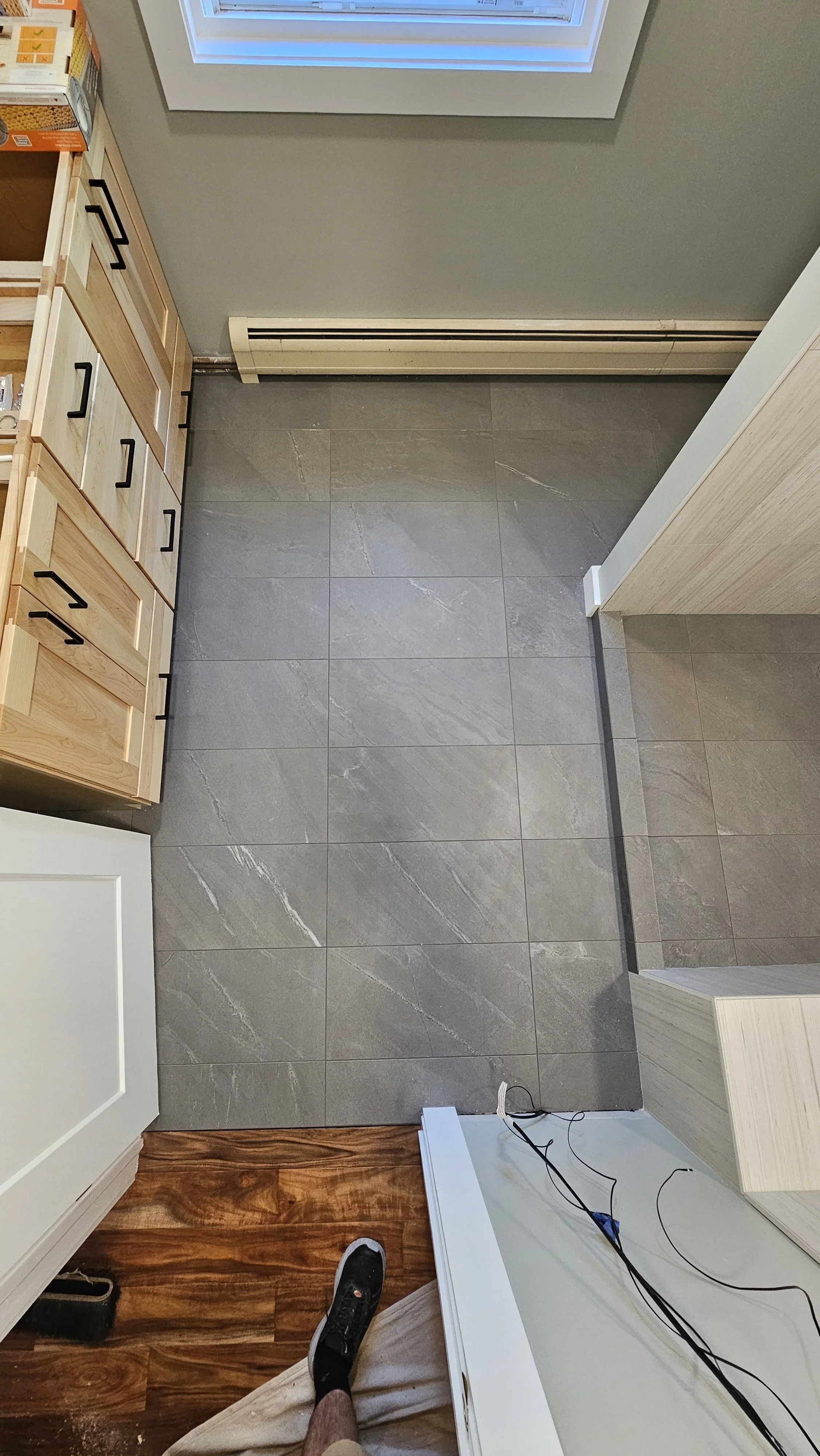 The photo shows a kitchen corner with a tiled gray floor, wooden cabinets, a window, and an electric baseboard heater along the wall. A person's foot wearing a black sneaker and a beige pant leg are visible, and some black electrical cords rest on a 