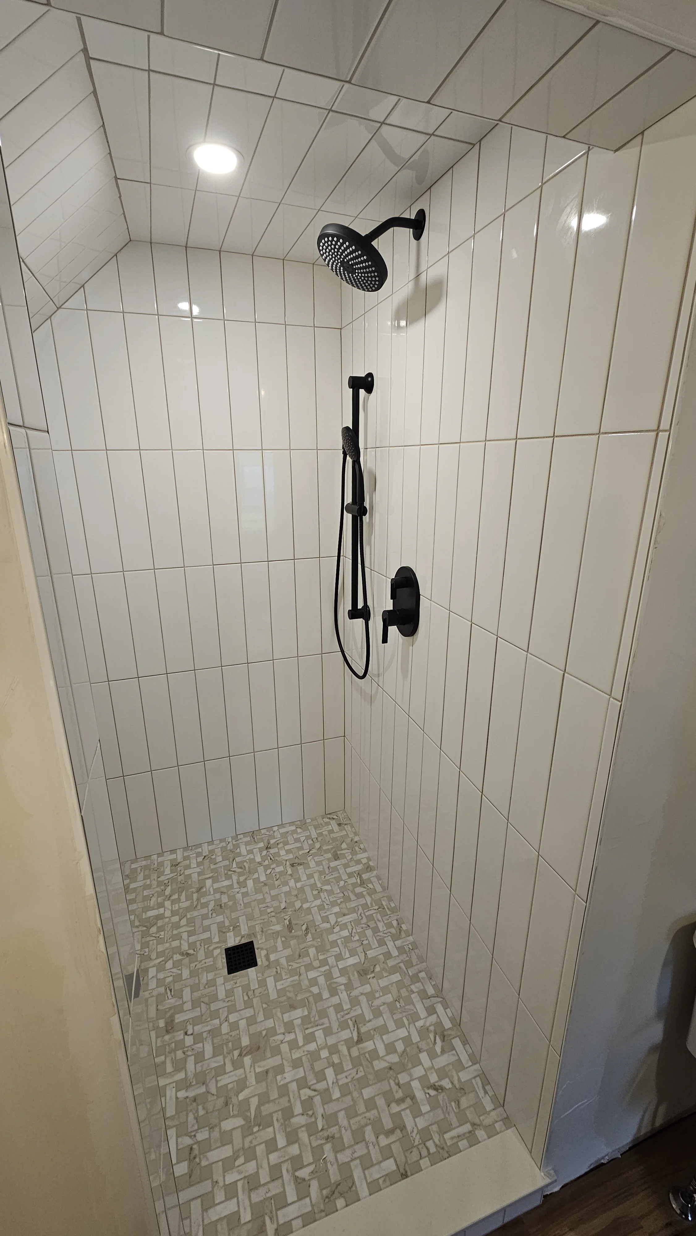 A small shower area with beige and white square tiles on the wall and a patterned tile floor. It has a black overhead rain showerhead, a black handheld showerhead, and a black control knob on the wall.