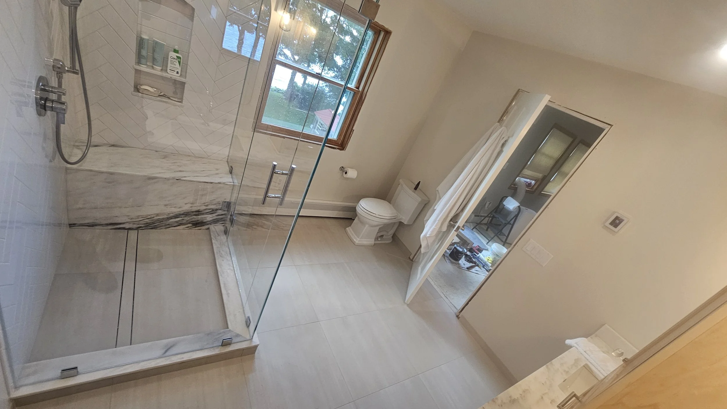 A bathroom with a glass shower enclosure, a toilet, a window, and a small mirror. The shower has white and gray veined marble panels, and there are toiletries on a shelf inside the shower. The room has beige walls and a beige floor, with a view of tr