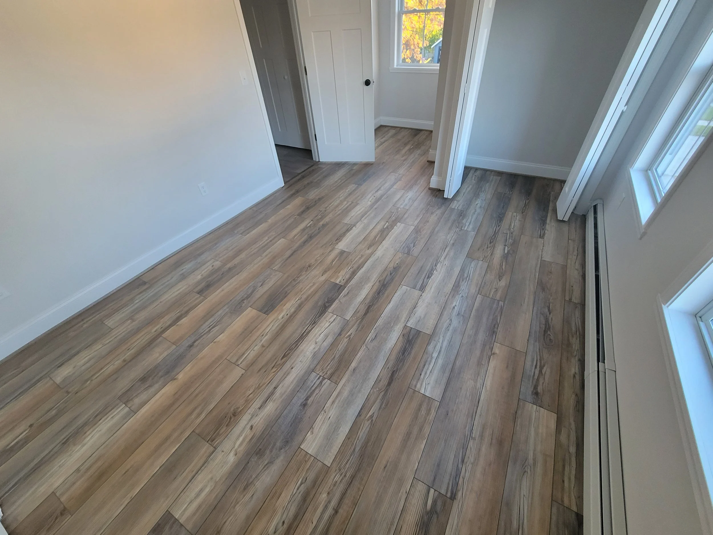Empty room with hardwood floors, white walls, a window showing autumn trees outside, and an open door leading to another room.