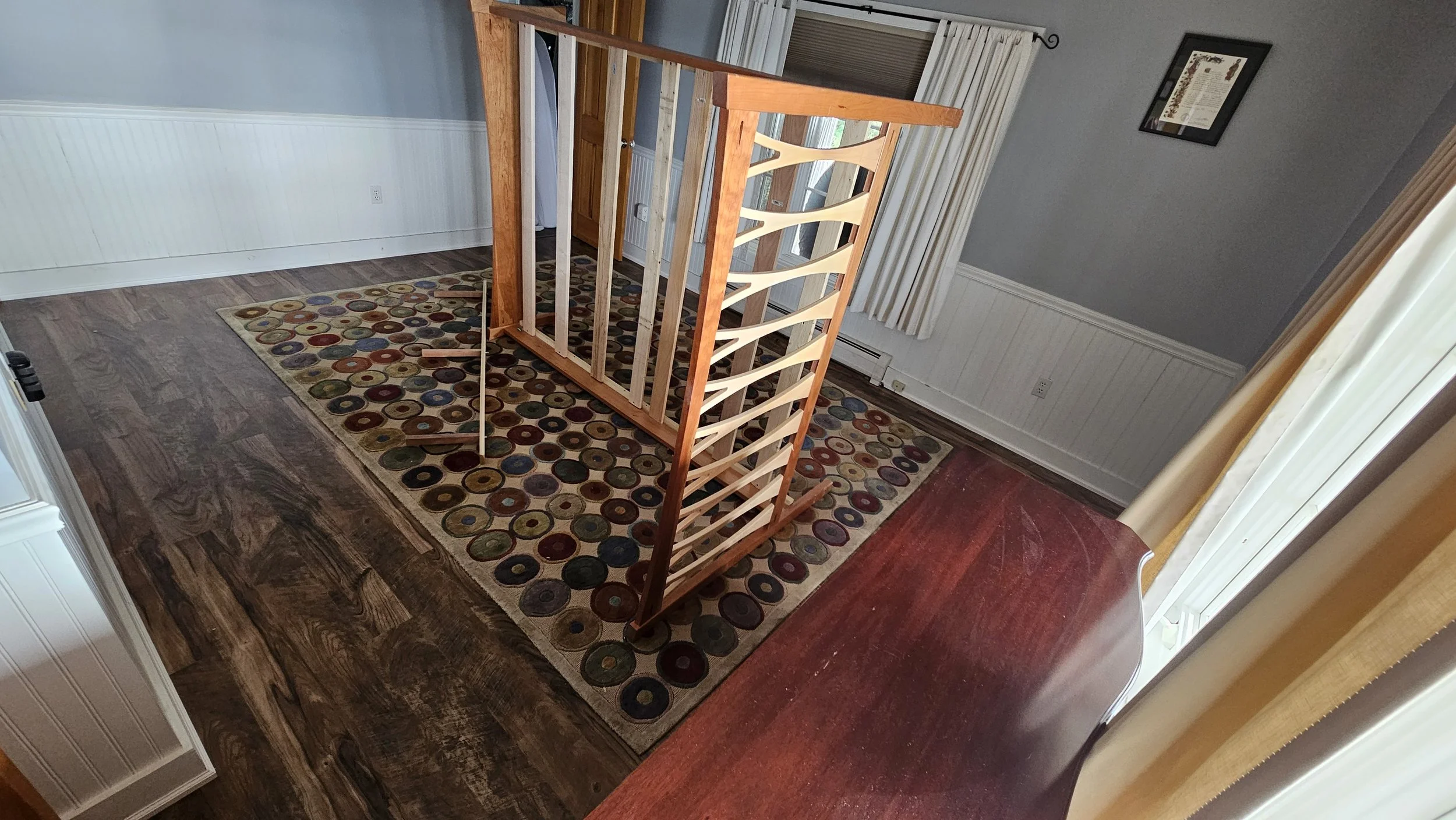 Inside a home, a wooden railing is placed on a colorful circular-patterned rug on a hardwood floor. A wall with light blue paneling, a framed picture, and a door are visible, with curtains hanging by a window.