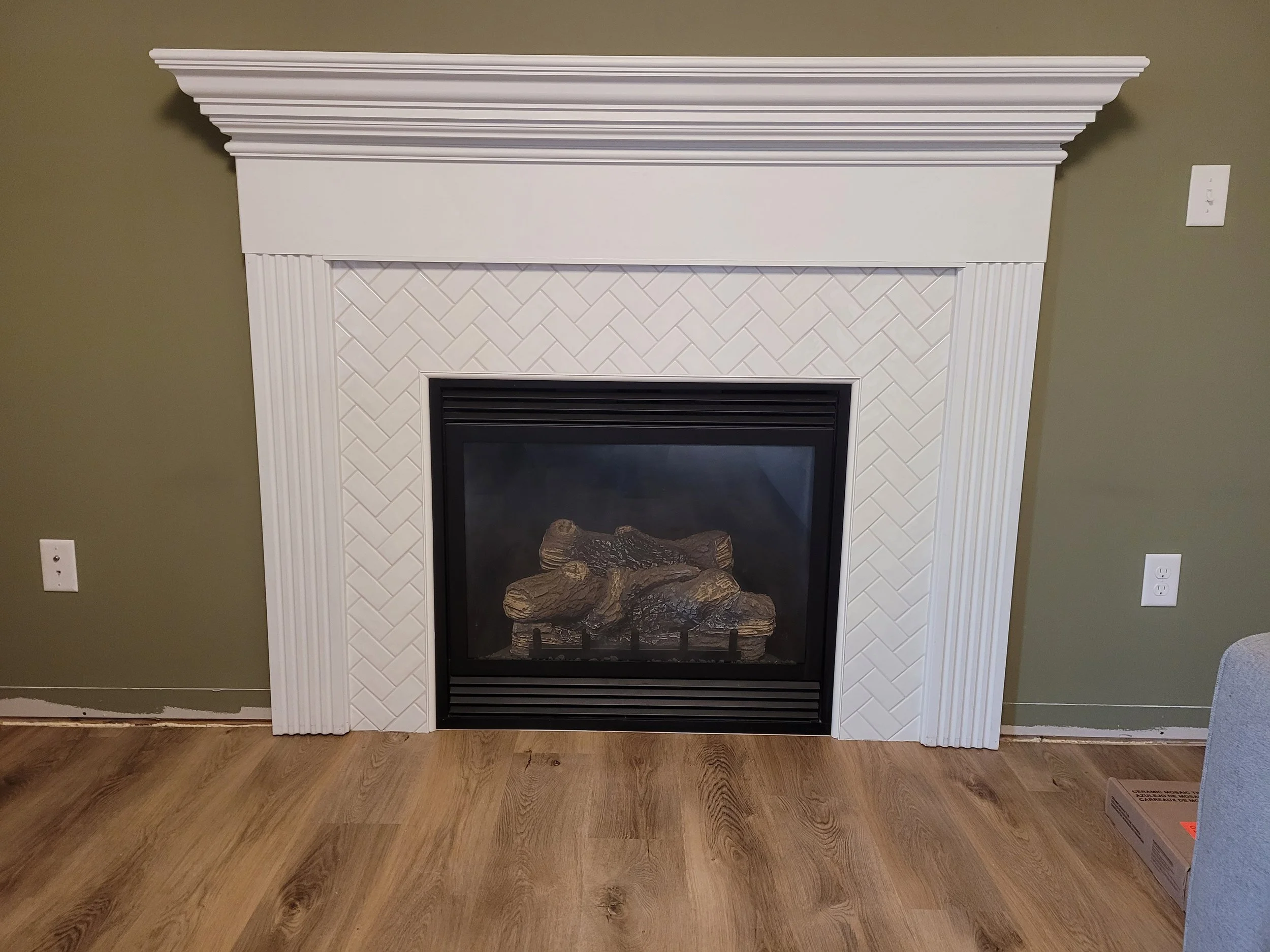 White fireplace mantel with a tile surround, featuring a herringbone pattern, and decorative molding on top. Inside the fireplace are logs with a grate.