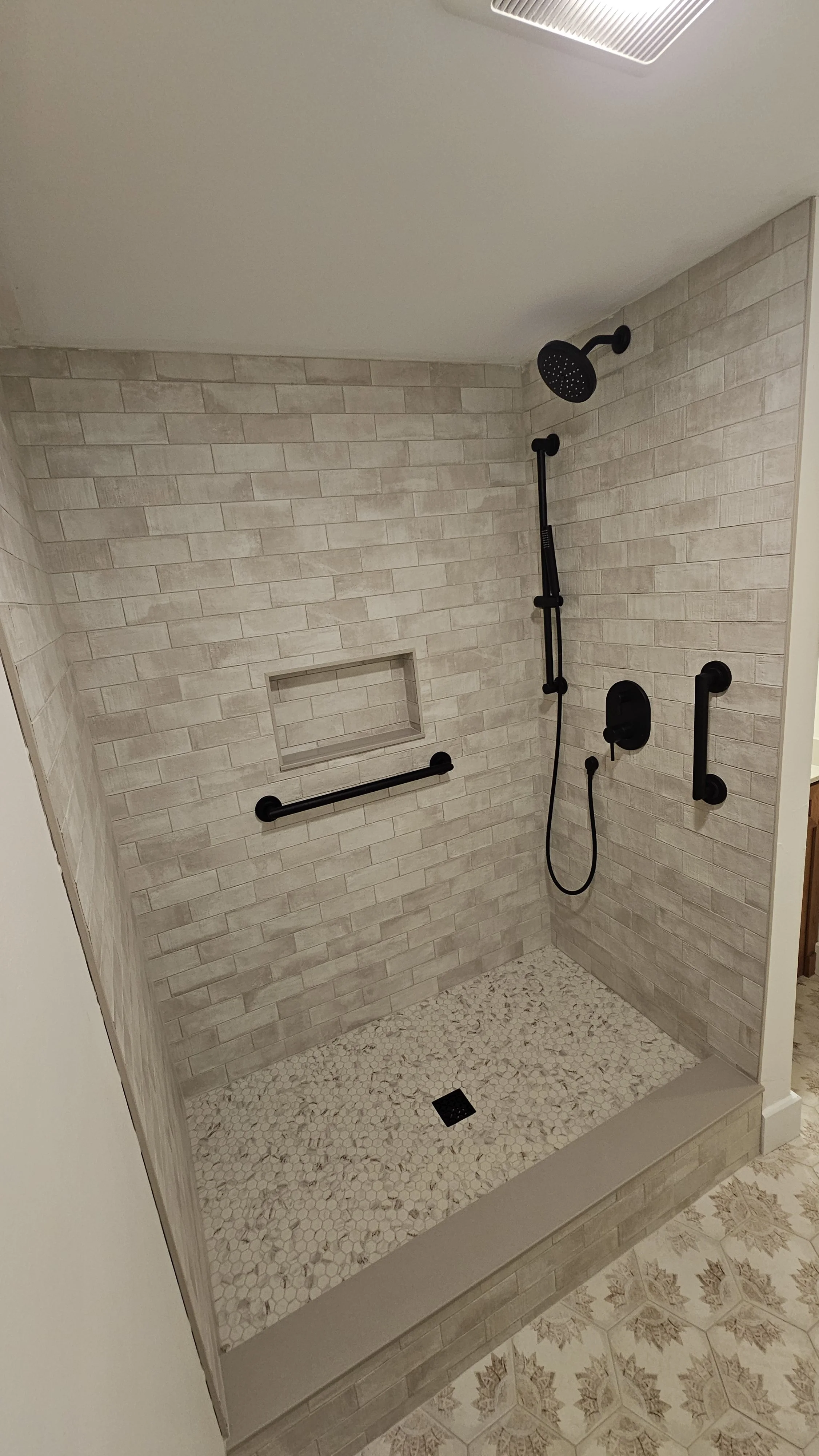 Shower with beige subway tile walls, a black rainfall showerhead, a handheld shower, two grab bars, a built-in niche, and pebble tile floor.