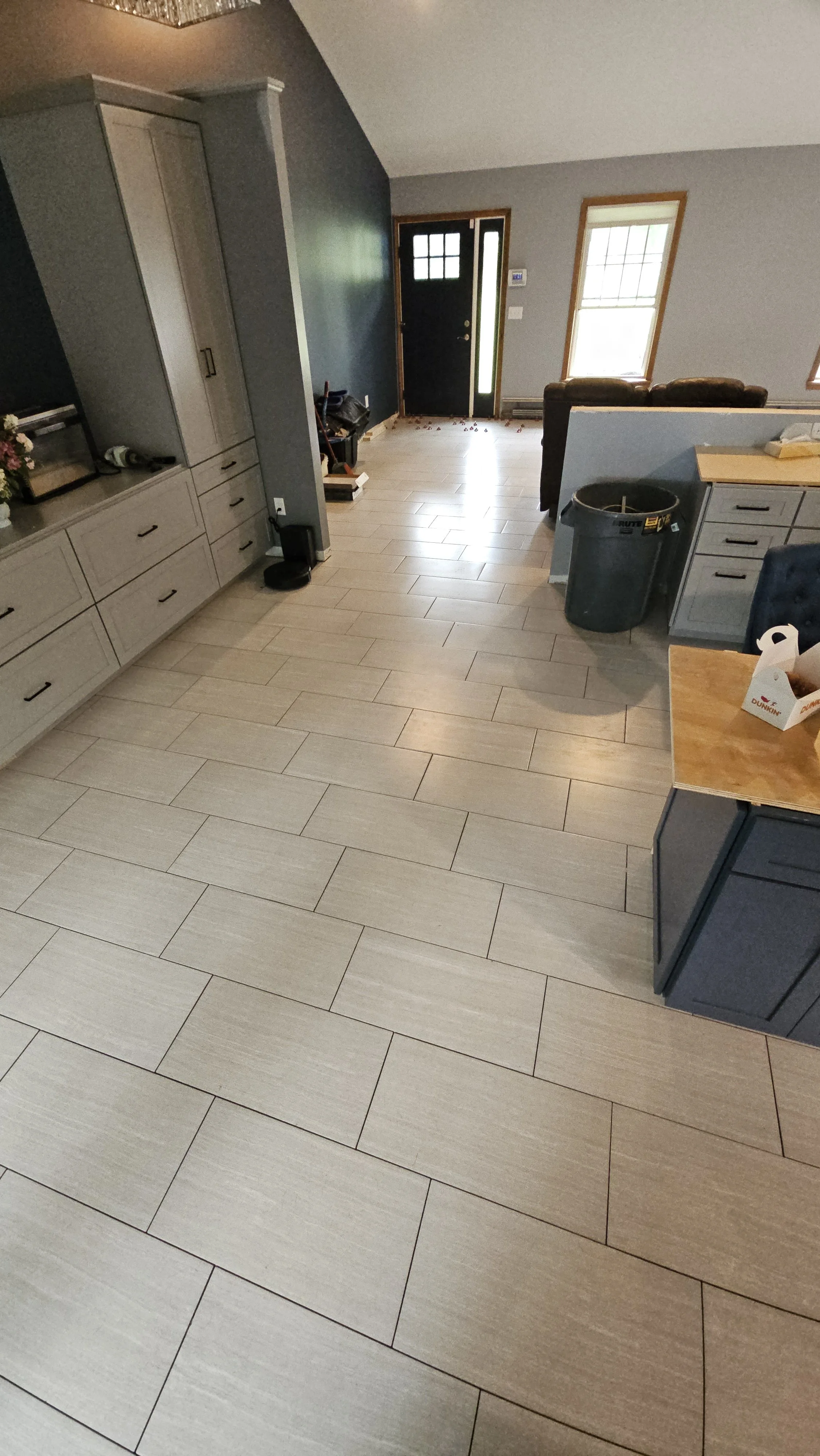 Interior view of a house entryway with light-colored wood floor tiles, a black front door with glass panel, and two windows. There are kitchen cabinets and appliances on the right, a trash can, and a living room with a sofa. The space appears to be i