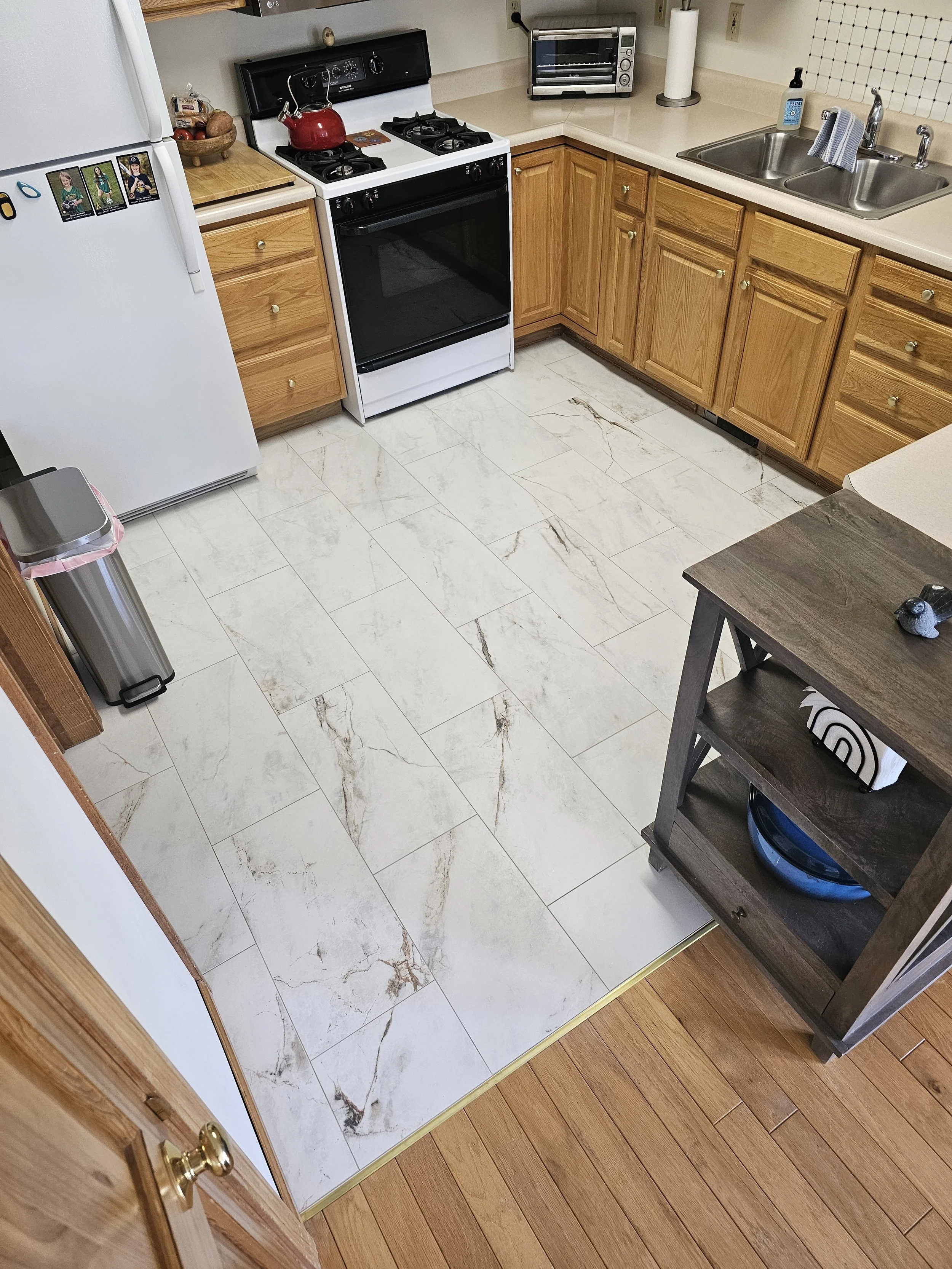 A kitchen with a white refrigerator on the left, a stove with a red kettle on top, a small countertop oven, a sink with a paper towel roll, and various small items. The flooring is a mix of tile and wood.