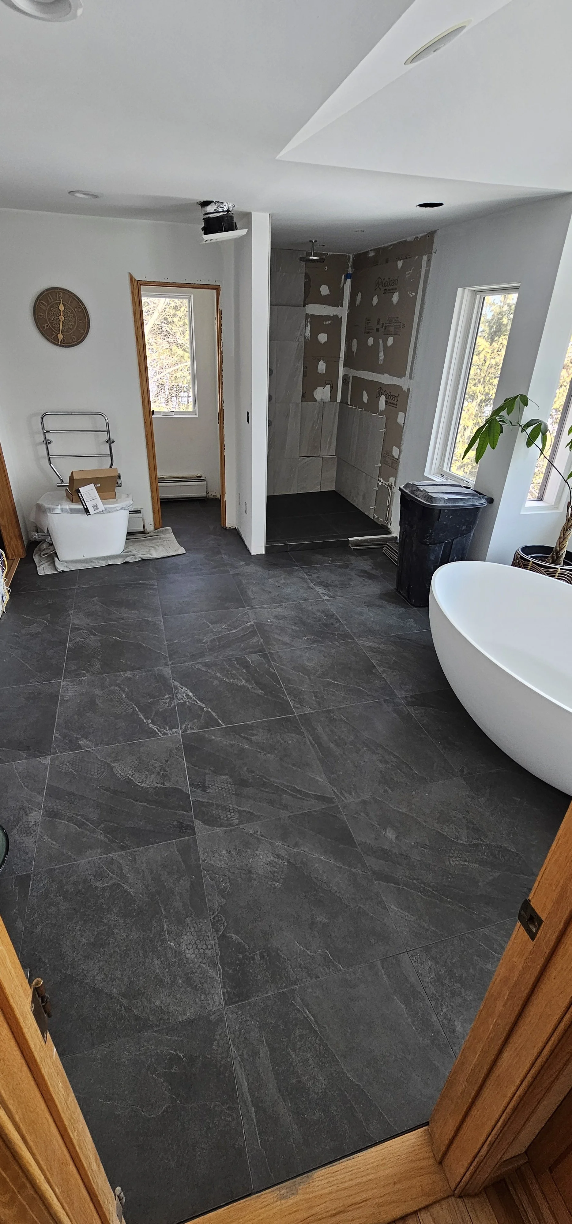 Bathroom under renovation with a black stone floor, an unfinished shower with tiling, a white freestanding bathtub, a window with trees outside, and construction materials placed around.