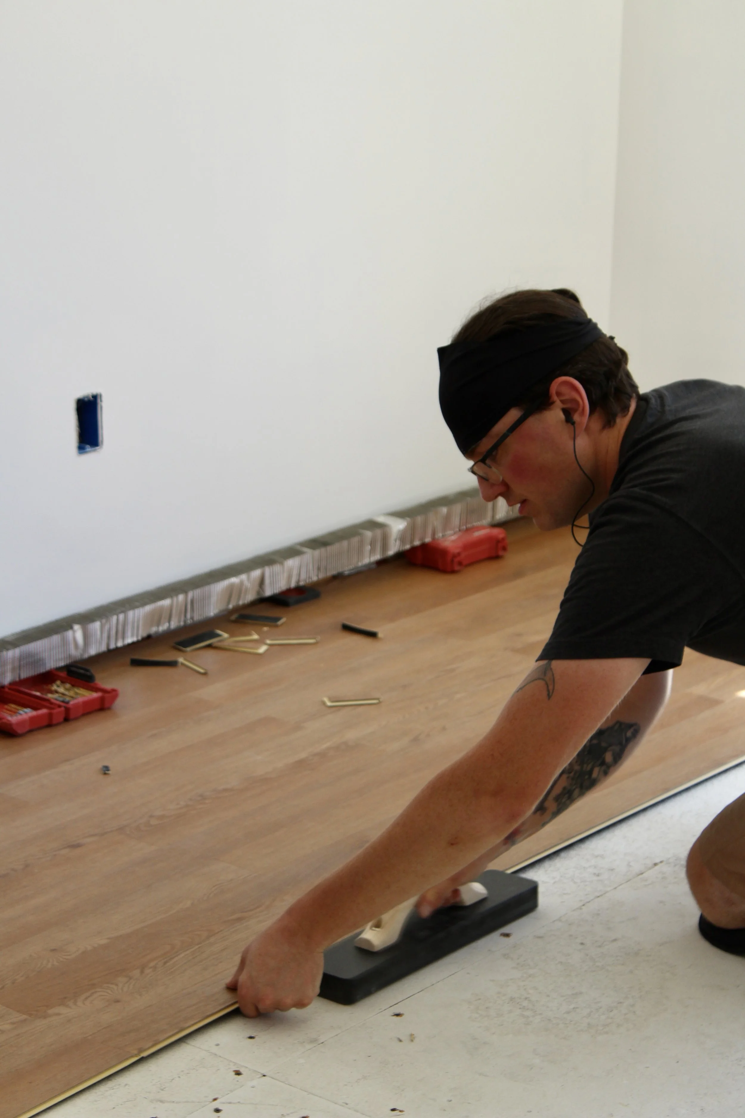 Person in black headwrap and glasses installing hardwood flooring, using a flooring tapping block and mallet, with tools and materials on the floor