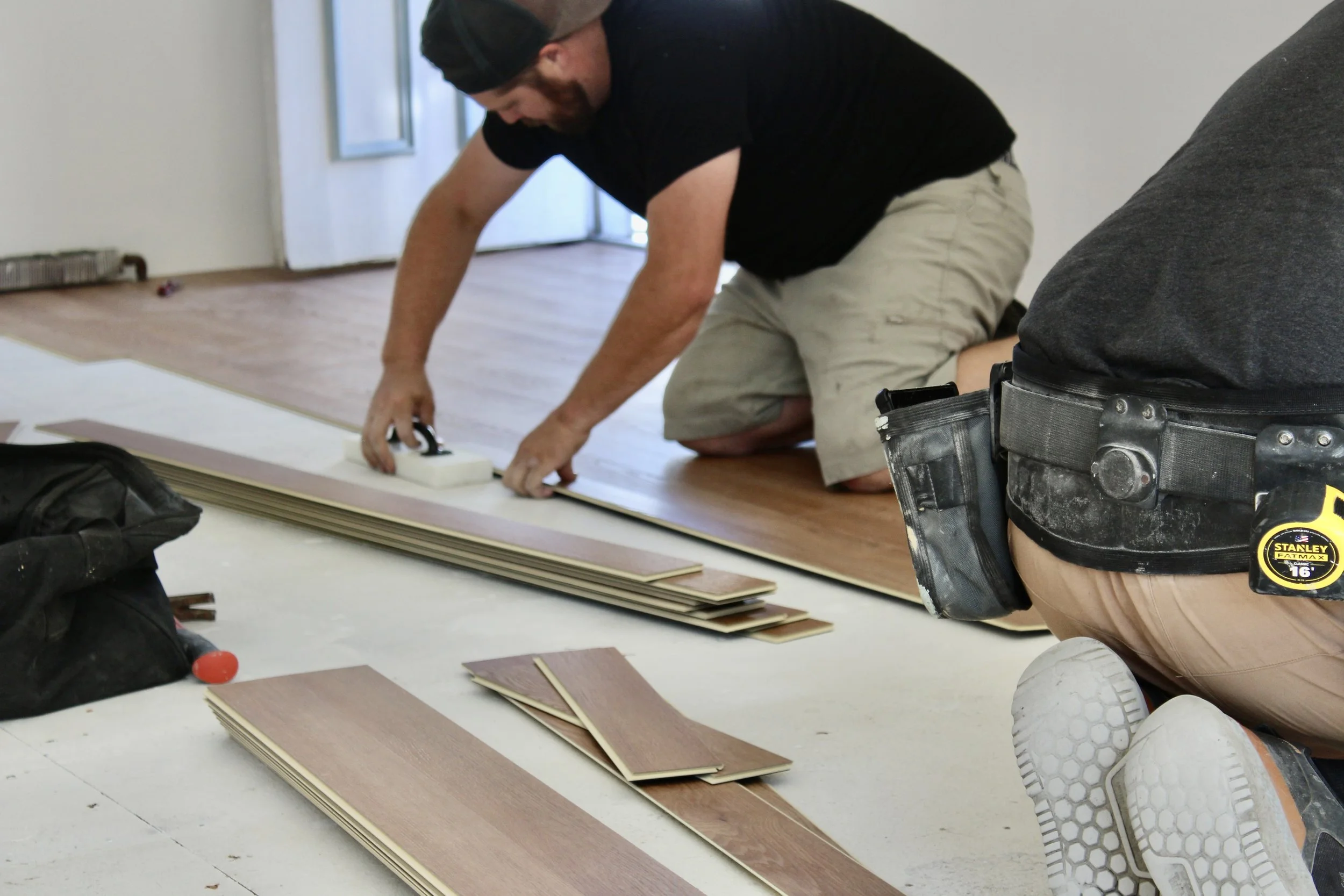 A man kneeling on the floor measuring wooden boards during a construction project indoors, with others partially visible nearby.
