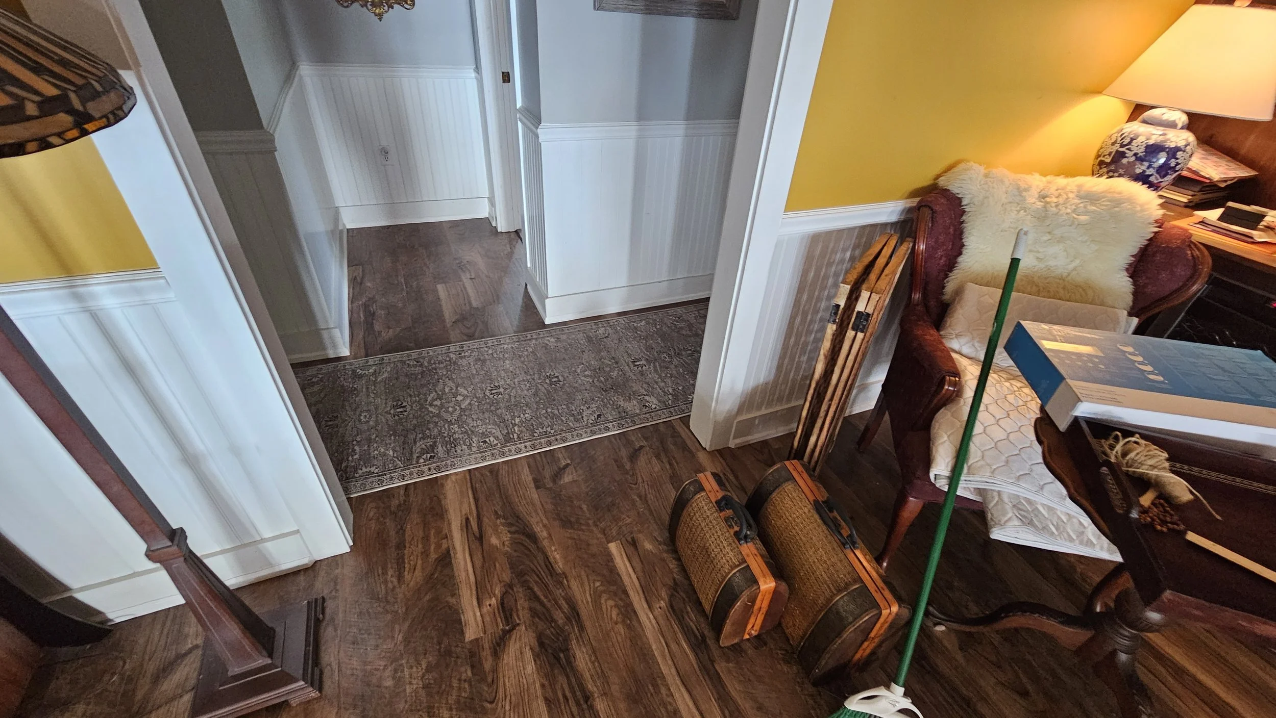 A cozy living room area with dark wood flooring, a vibrant yellow wall, a small beige and white rug, a wooden table with a white quilted cloth, various items including a lamp, books, and a blanket, and cleaning supplies like a mop and containers unde
