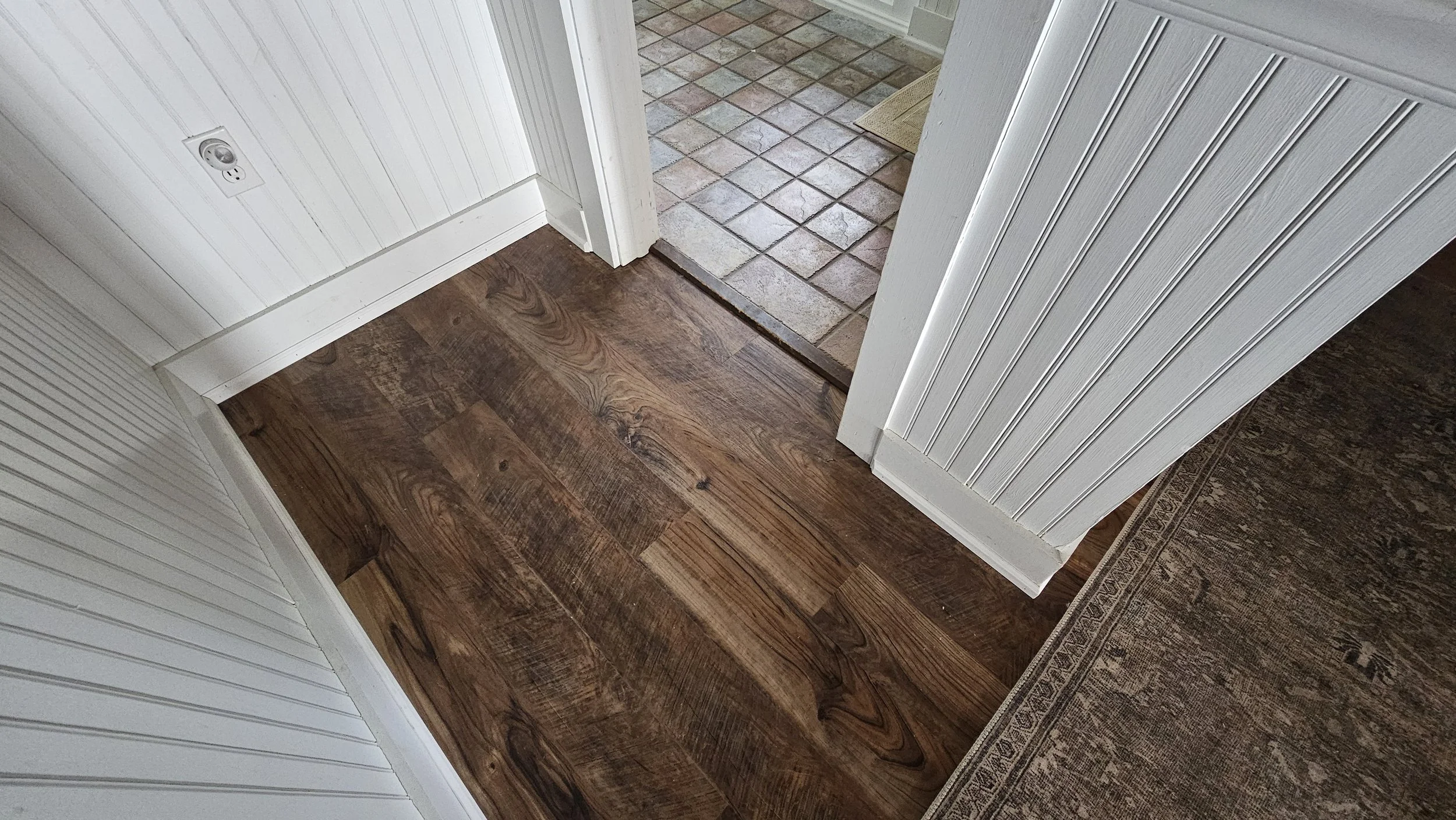 Interior corner of a room showing wooden flooring, a tiled entryway, and white paneled walls with trim.