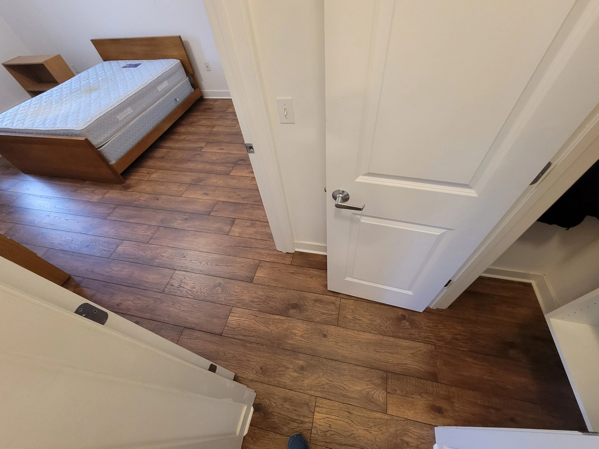 View of an open white door leading into a room with wooden flooring, a bed with a mattress and a small wooden nightstand.