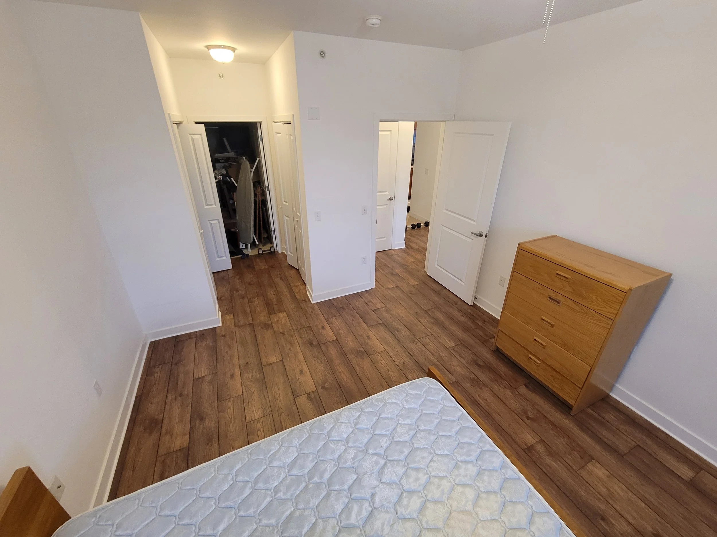 Empty bedroom with white walls, wood flooring, a wooden dresser, and an open closet with various items inside. Part of a mattress is visible in the foreground.