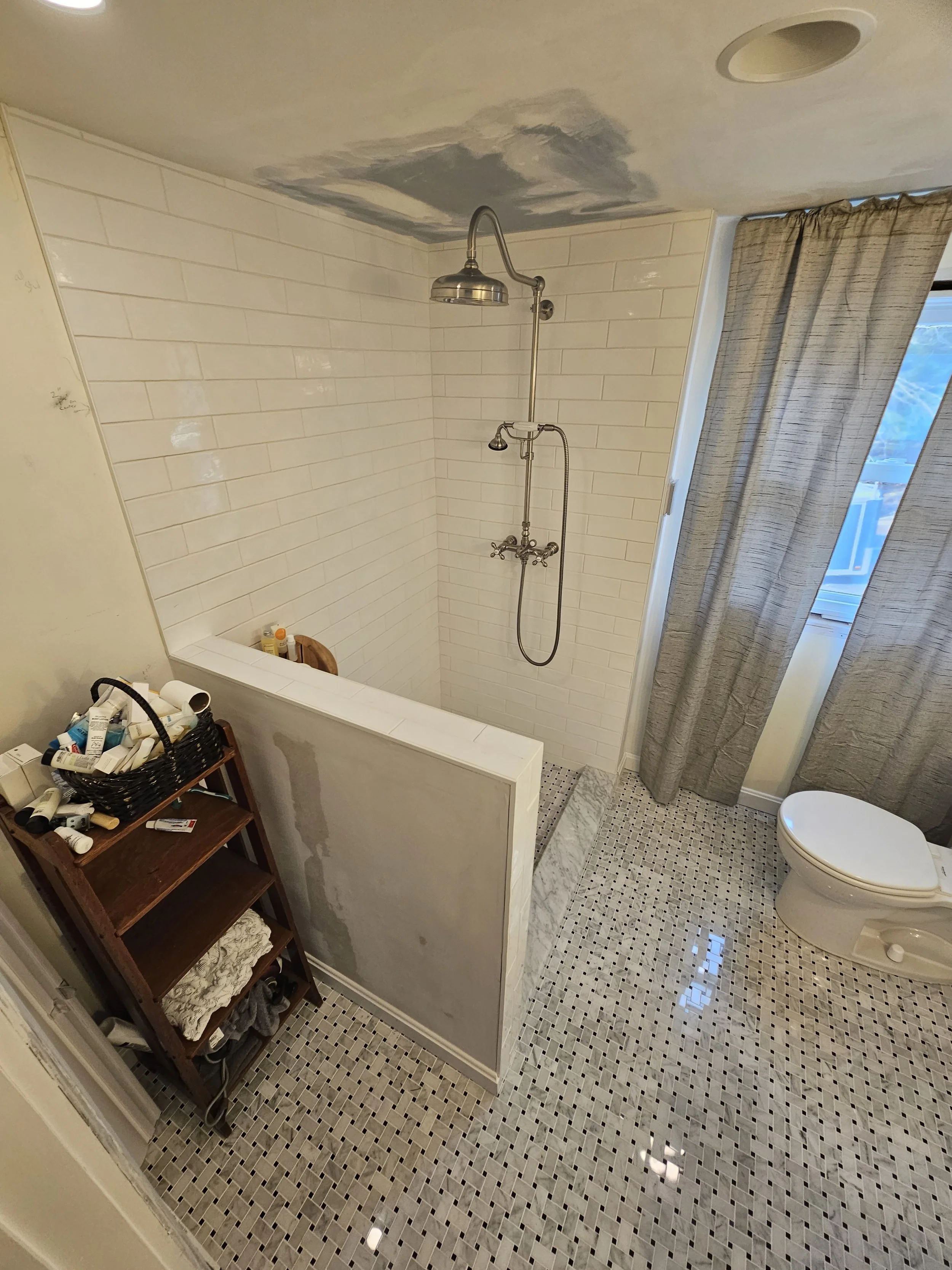 A partially renovated bathroom with a black and white mosaic floor, a white tiled shower area with an overhead showerhead, and a toilet. There is a wooden shelf holding toiletries and a half-wall separating the shower from the rest of the bathroom. A
