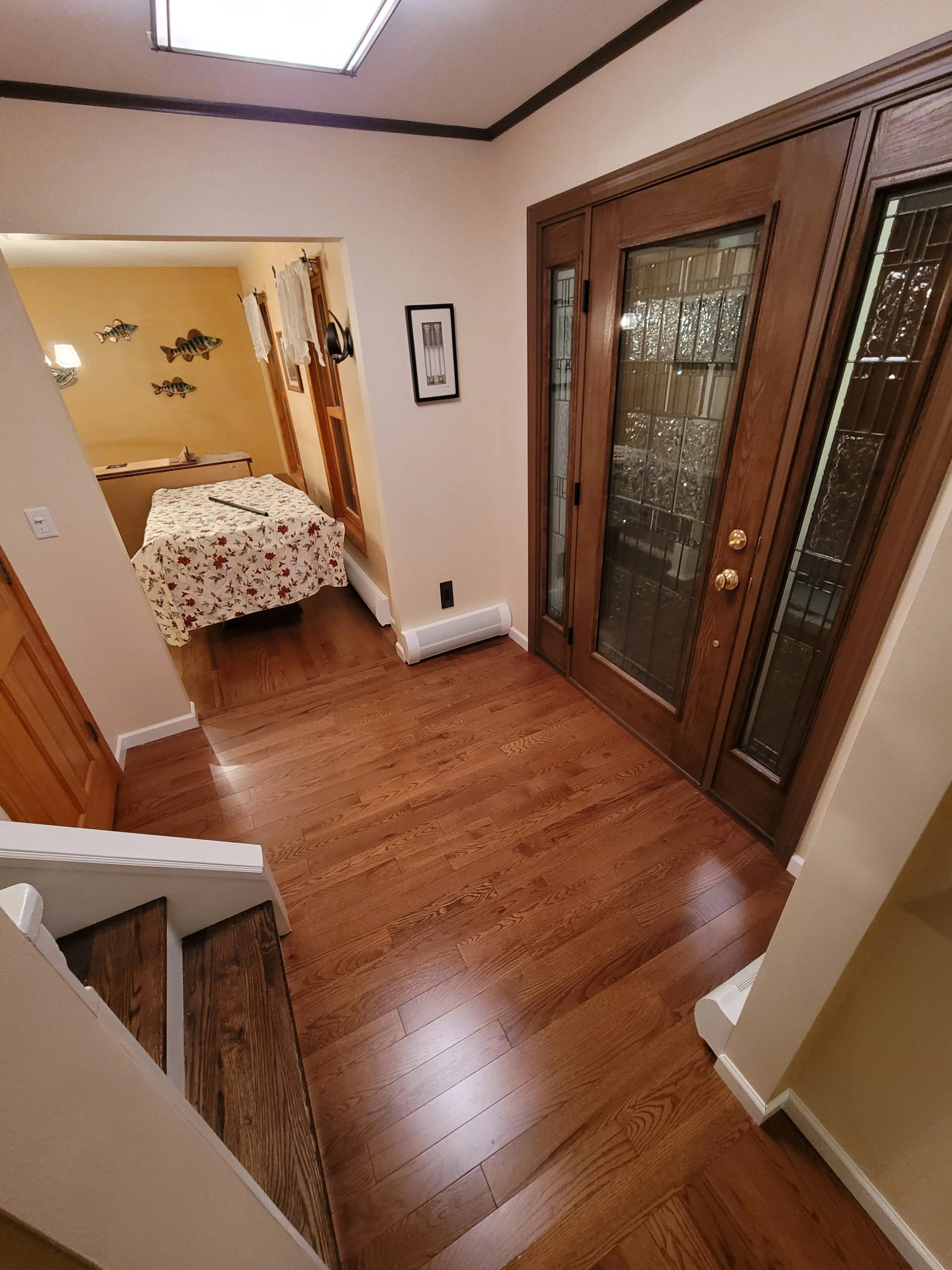 A hallway with hardwood floors, beige walls, and white baseboards. There is a wooden cabinet with glass doors on the right and a framed picture on the wall. At the far end, a small room with yellow walls is visible, decorated with fish wall art and a