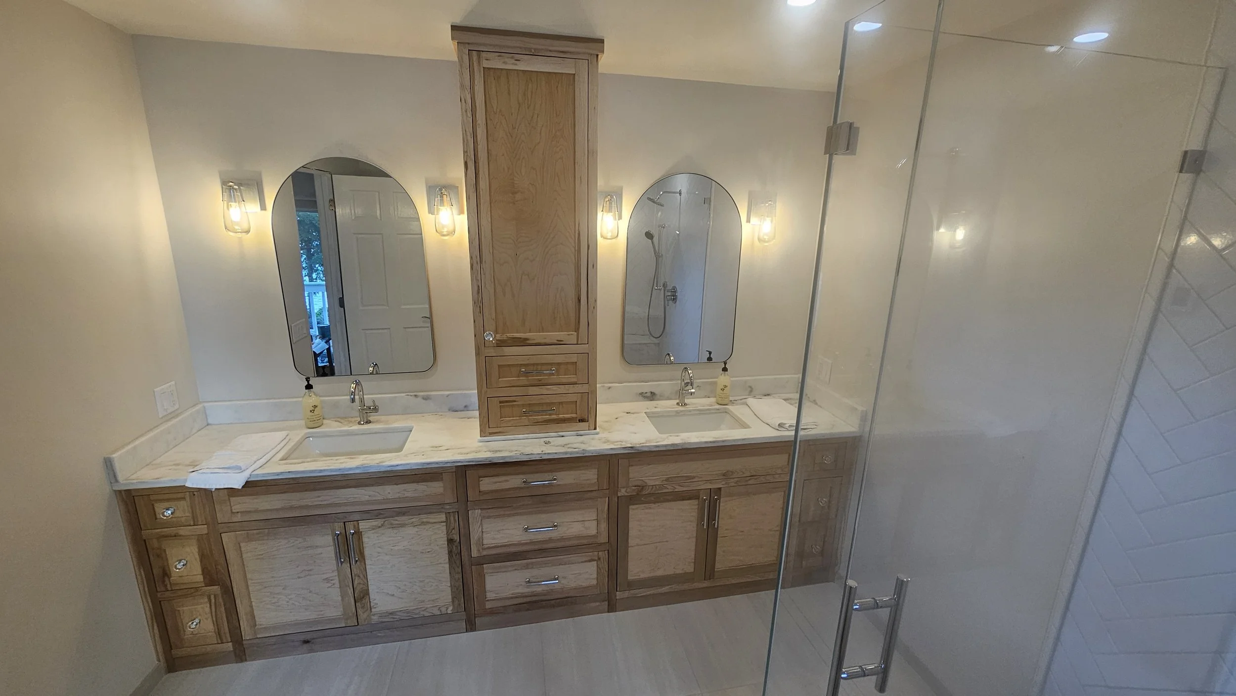 Bathroom with double sinks, mirrors, wooden cabinets, marble countertop, towel, soap dispenser, and shower with glass door.