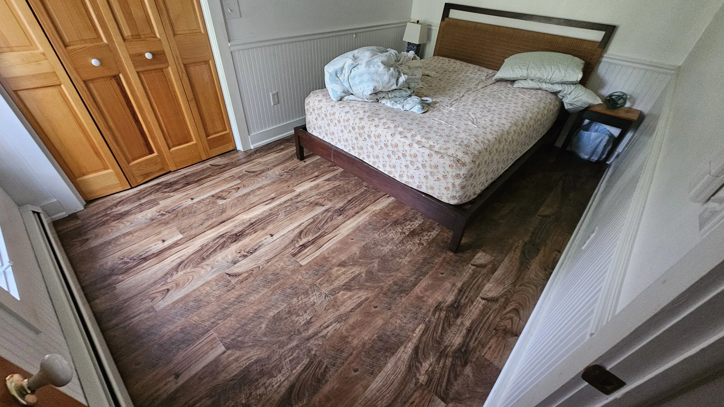 A bedroom with a bed that has a floral mattress cover and two pillows, one on each side. There is a nightstand with a lamp and a small object on it. The floor is wooden, with a closet with wooden doors on the left side.