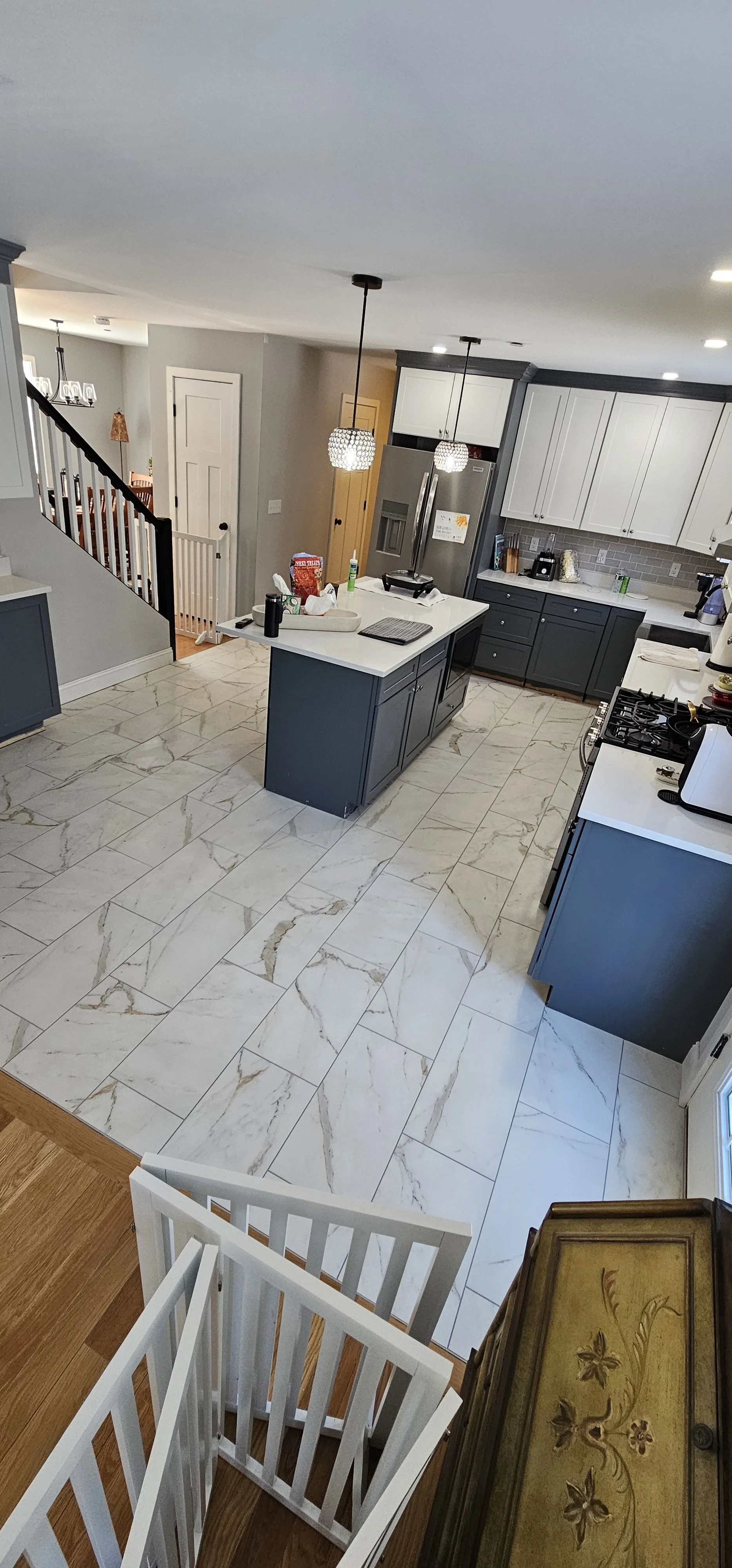Modern kitchen with white marble floor tiles, gray cabinetry, and a kitchen island. There are two pendant lights hanging above the island, a refrigerator, stovetop, and various kitchen appliances.