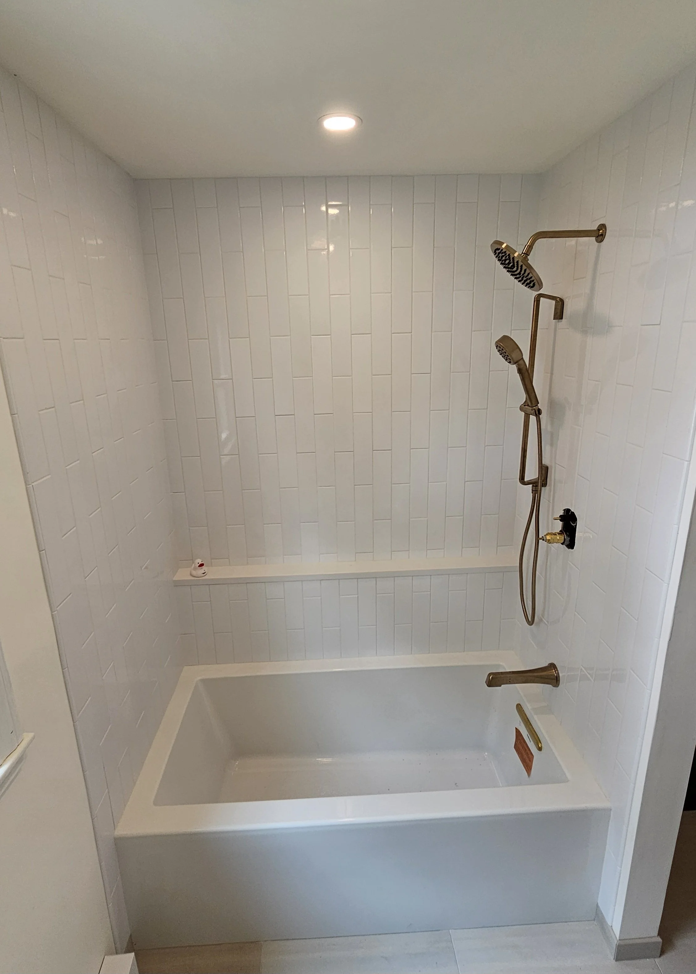 A bathroom shower with white tile walls, a built-in bathtub, and gold-colored fixtures including a showerhead, a handheld shower, and a grab bar.