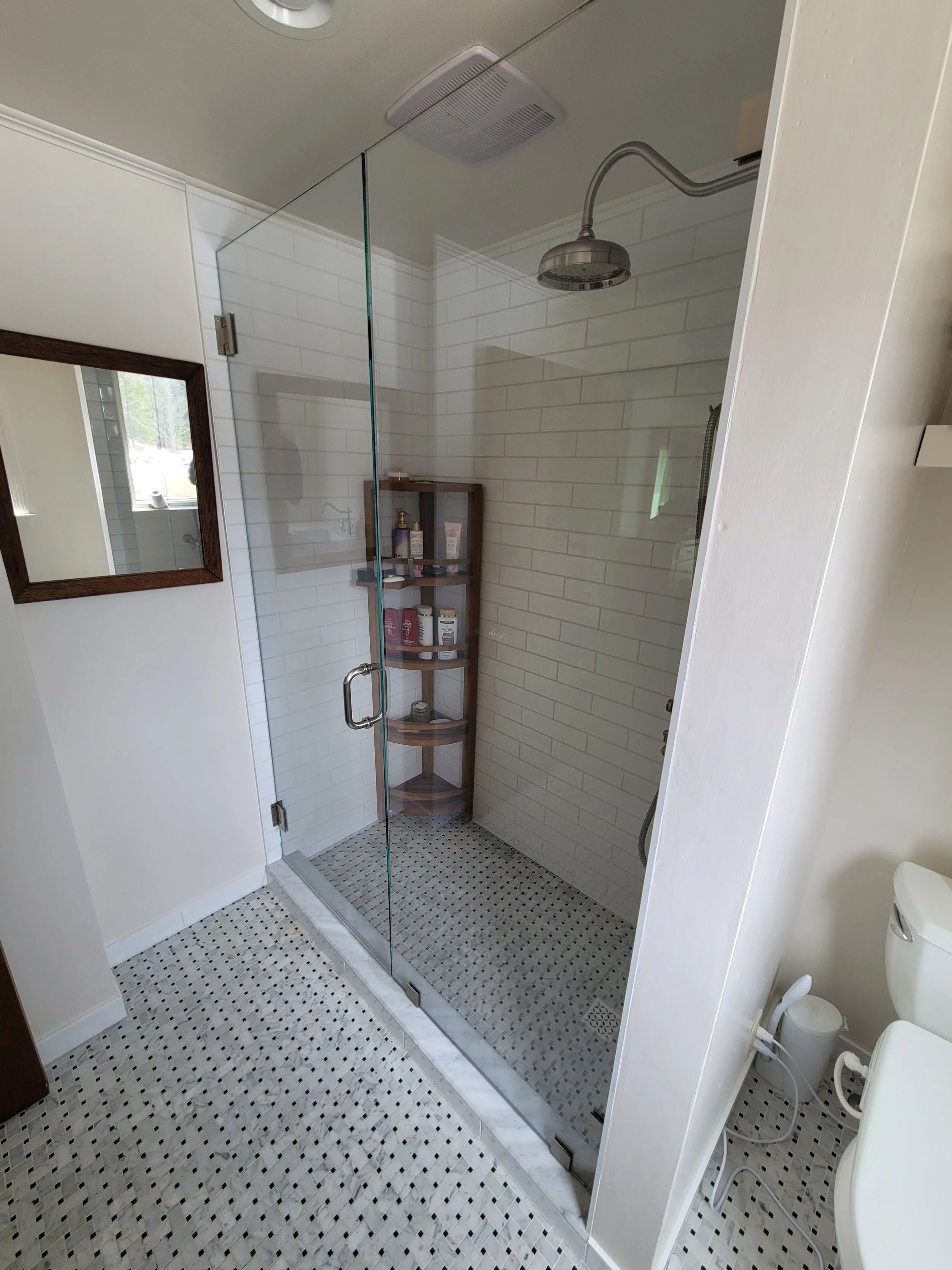 Shower with glass door, white subway wall tiles, a niche with toiletries, black and white mosaic floor tiles, a small window, ceiling vent, and showerhead.