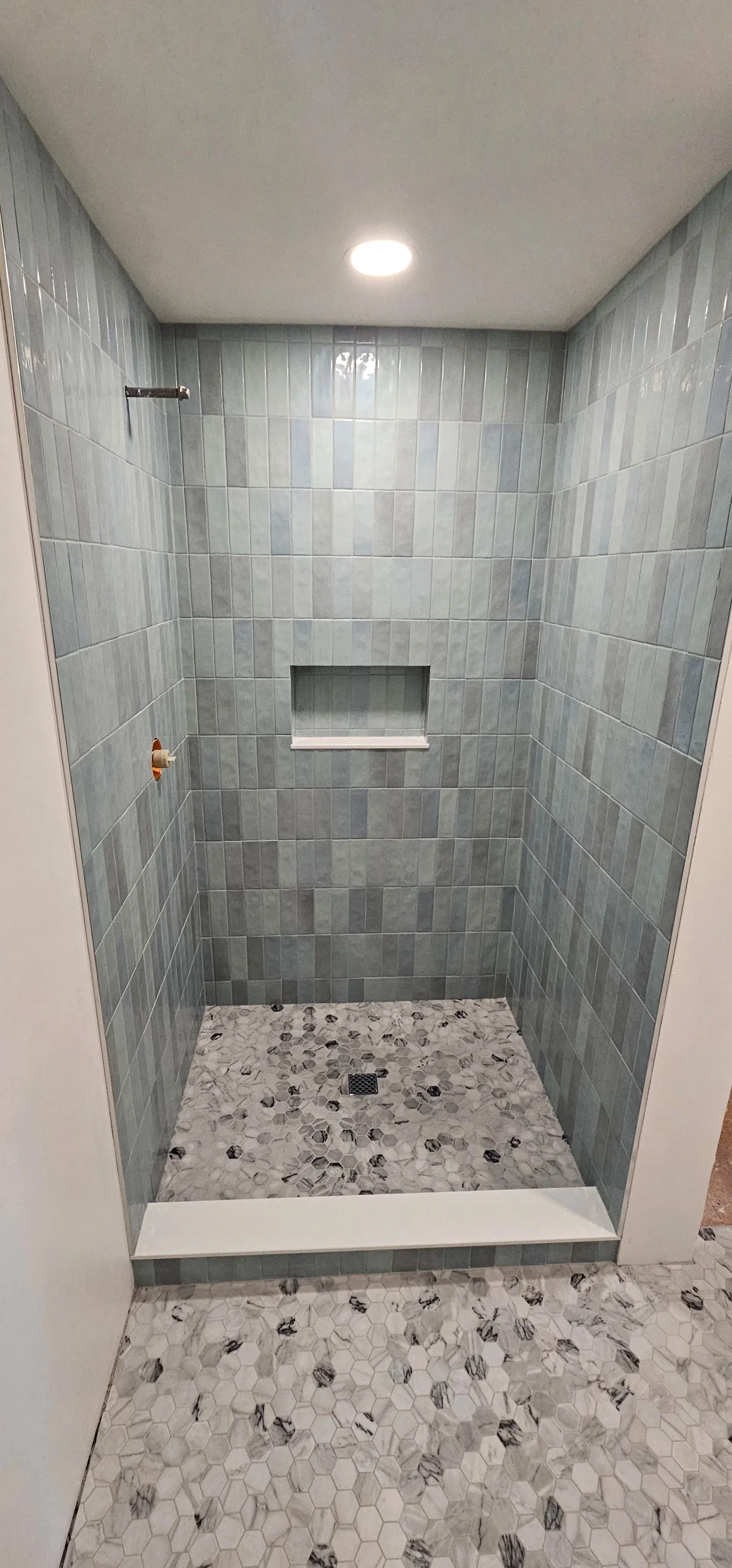 Empty walk-in shower with blue-gray tiled walls, a built-in niche for toiletries, a drain on the patterned marble floor, and a ceiling light.
