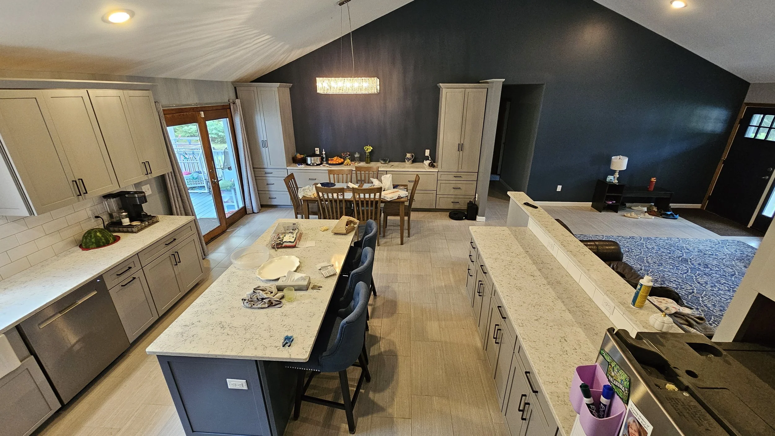 A kitchen with beige cabinets, a large white marble island with blue chairs, and a dining area with a wooden table and chairs. The back wall is painted dark, and there are sliding glass doors leading outside. The countertop has dishes and food items,