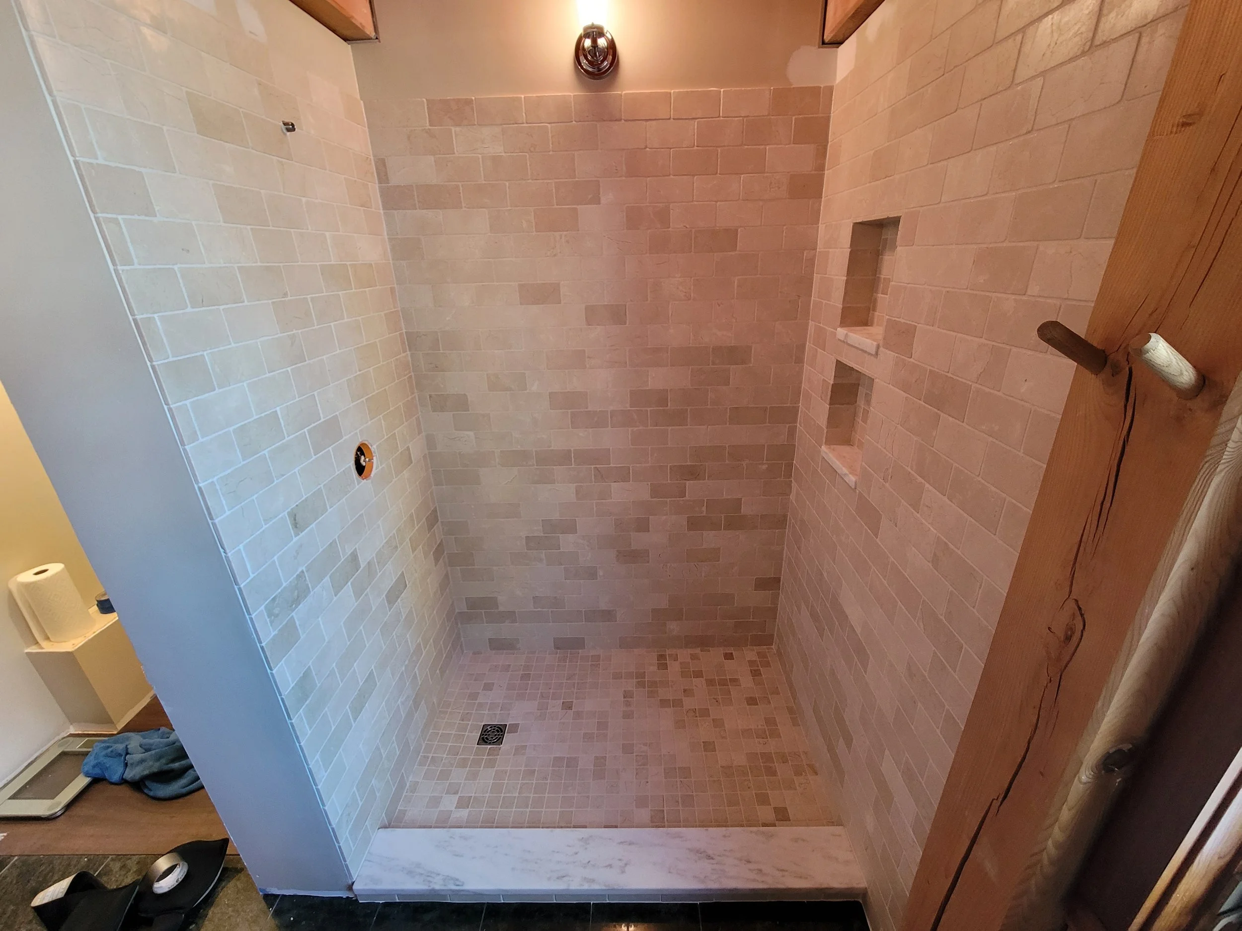 An unfinished walk-in shower with beige tile walls and small matching tile floor. There are built-in shelves and a drain on the floor. A hole in the wall is visible, likely for plumbing, and a vent is at the top of the wall. The edge of a wooden coun