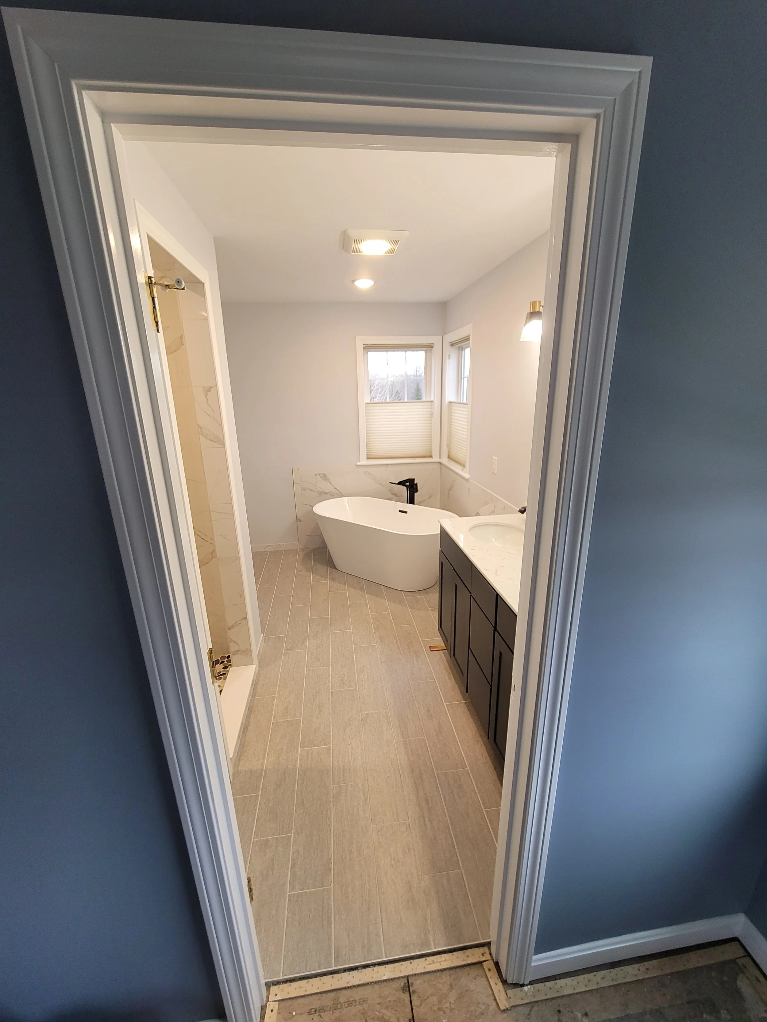A bathroom with a free-standing bathtub, a dark vanity with a white marble top, a window with blinds, a shower with a glass door, and tiled floors.