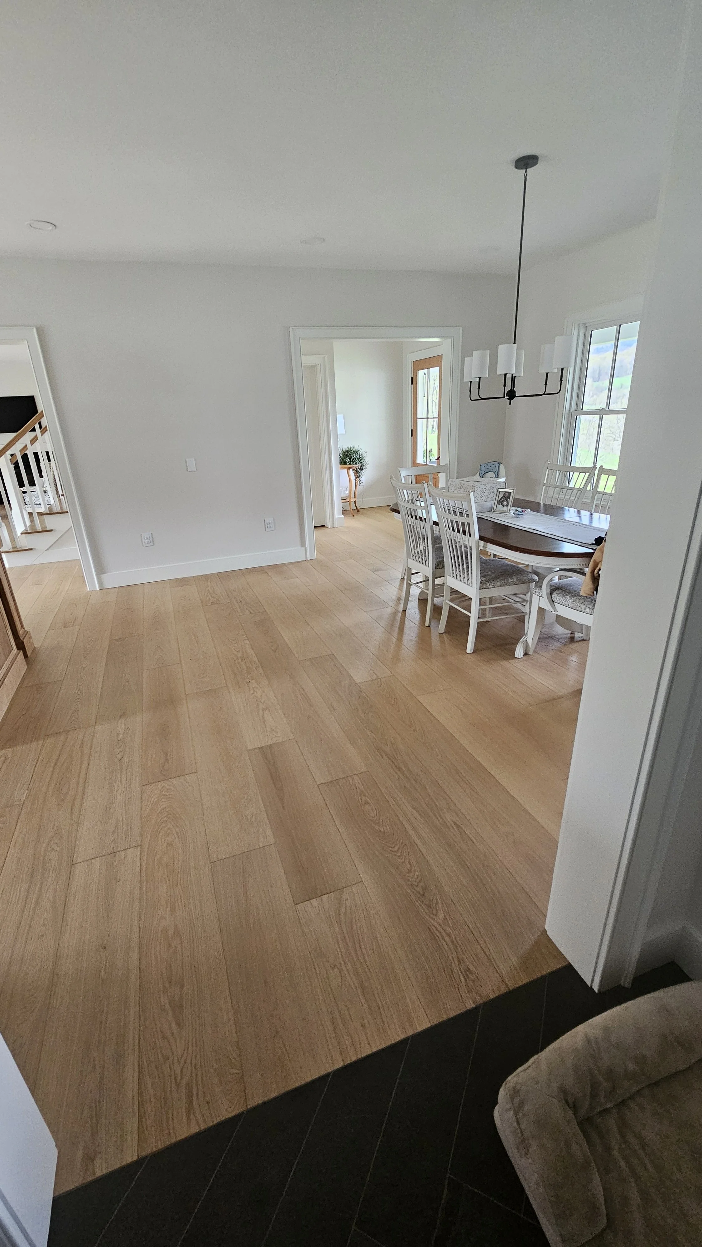 View of a dining room with a wooden table, white chairs, and a black chandelier. There are large windows and hardwood flooring.