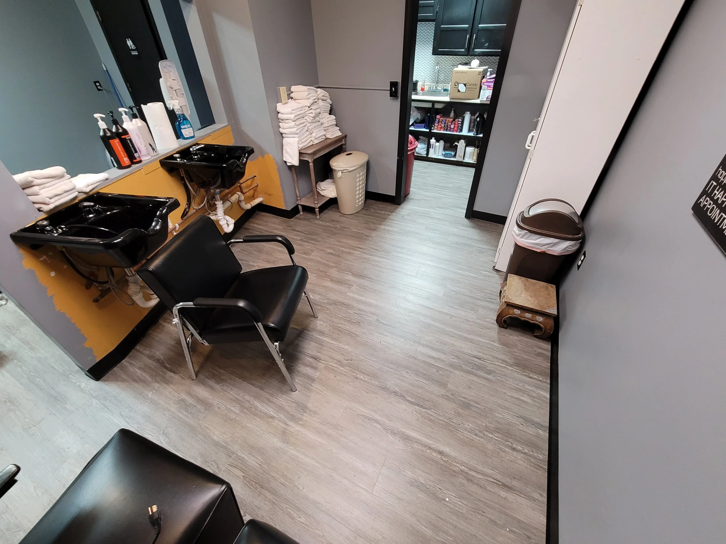 A small room with a black salon chair, a sink, and cleaning supplies on the window sill. There are towels folded on a shelf, a laundry basket, and a cabinet with personal care products inside. The walls are painted gray and there is wood flooring.