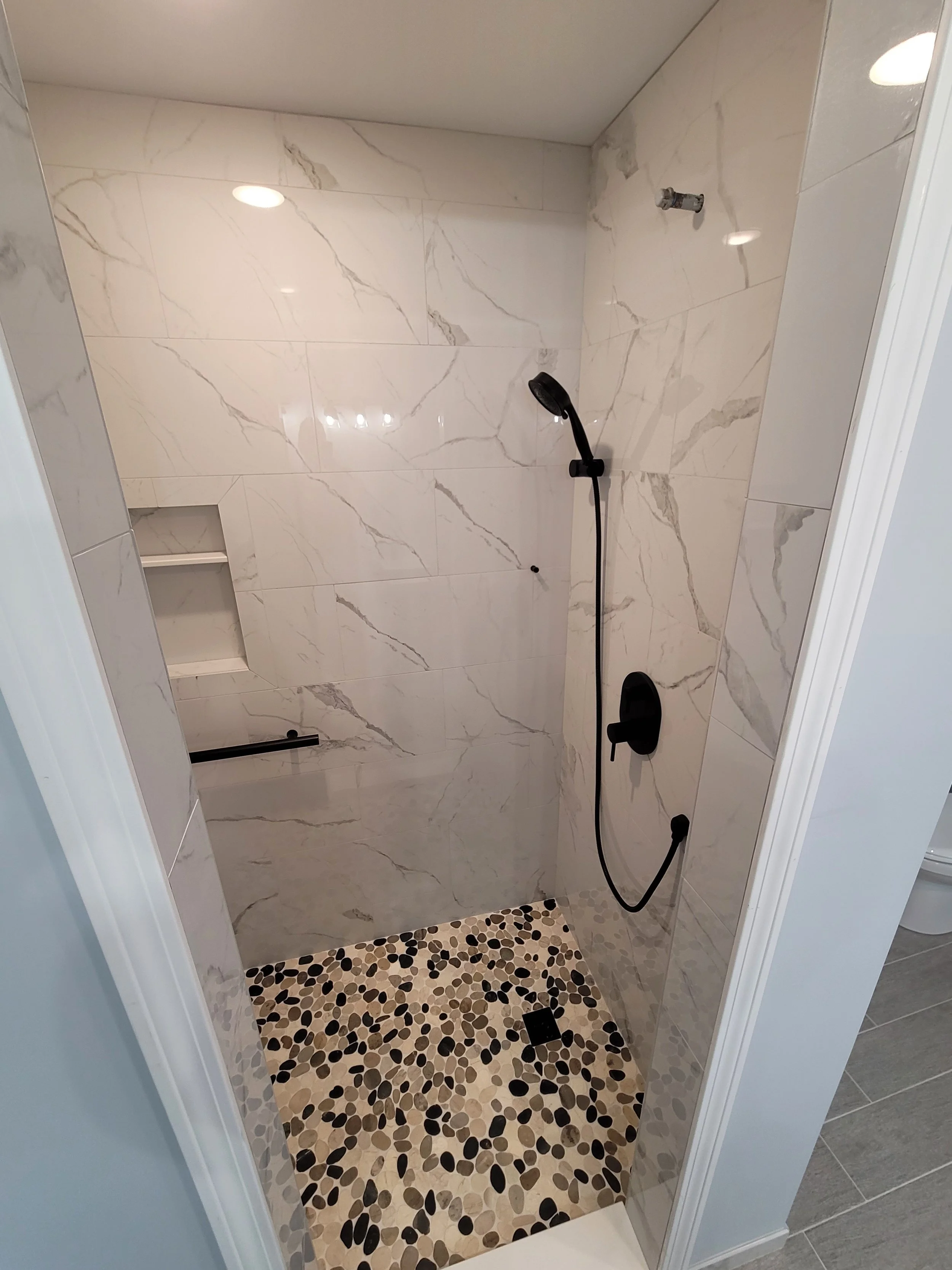 A walk-in shower with marble walls, a pebble tile floor, a black showerhead, and a built-in wall niche for toiletries.