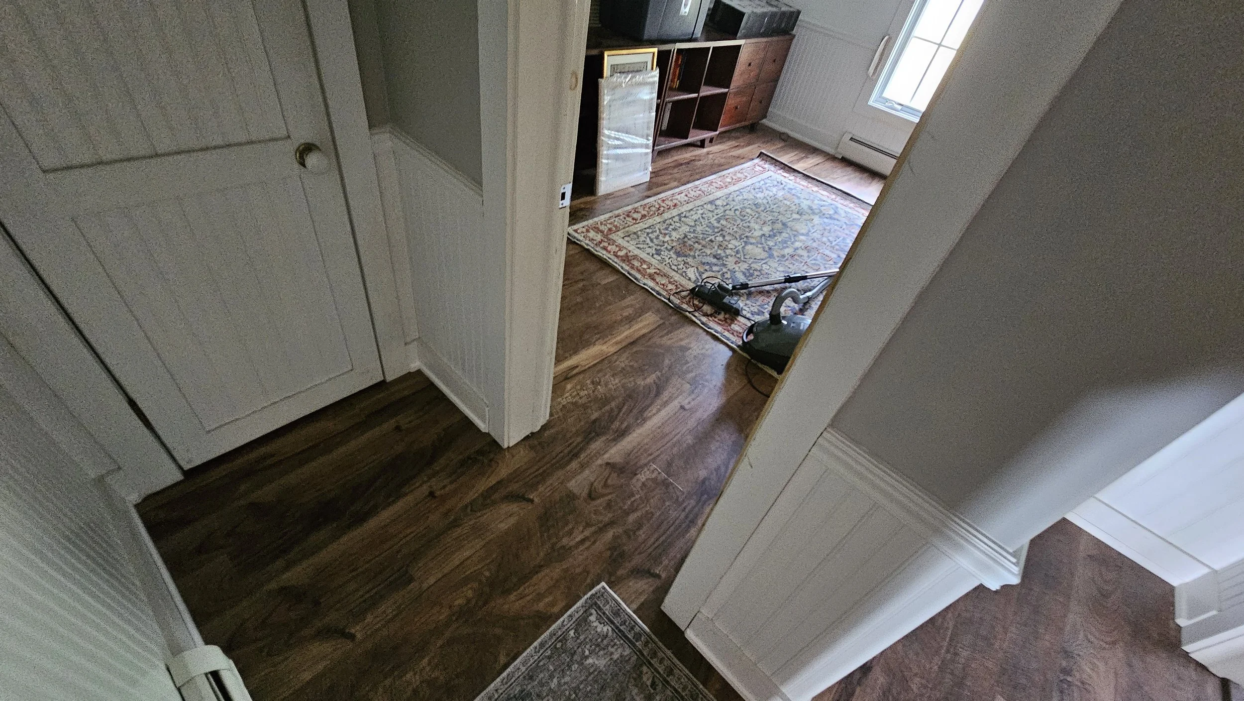 View of a room with hardwood flooring, a patterned rug, a vacuum cleaner, a media cabinet, a window, and a radiator.