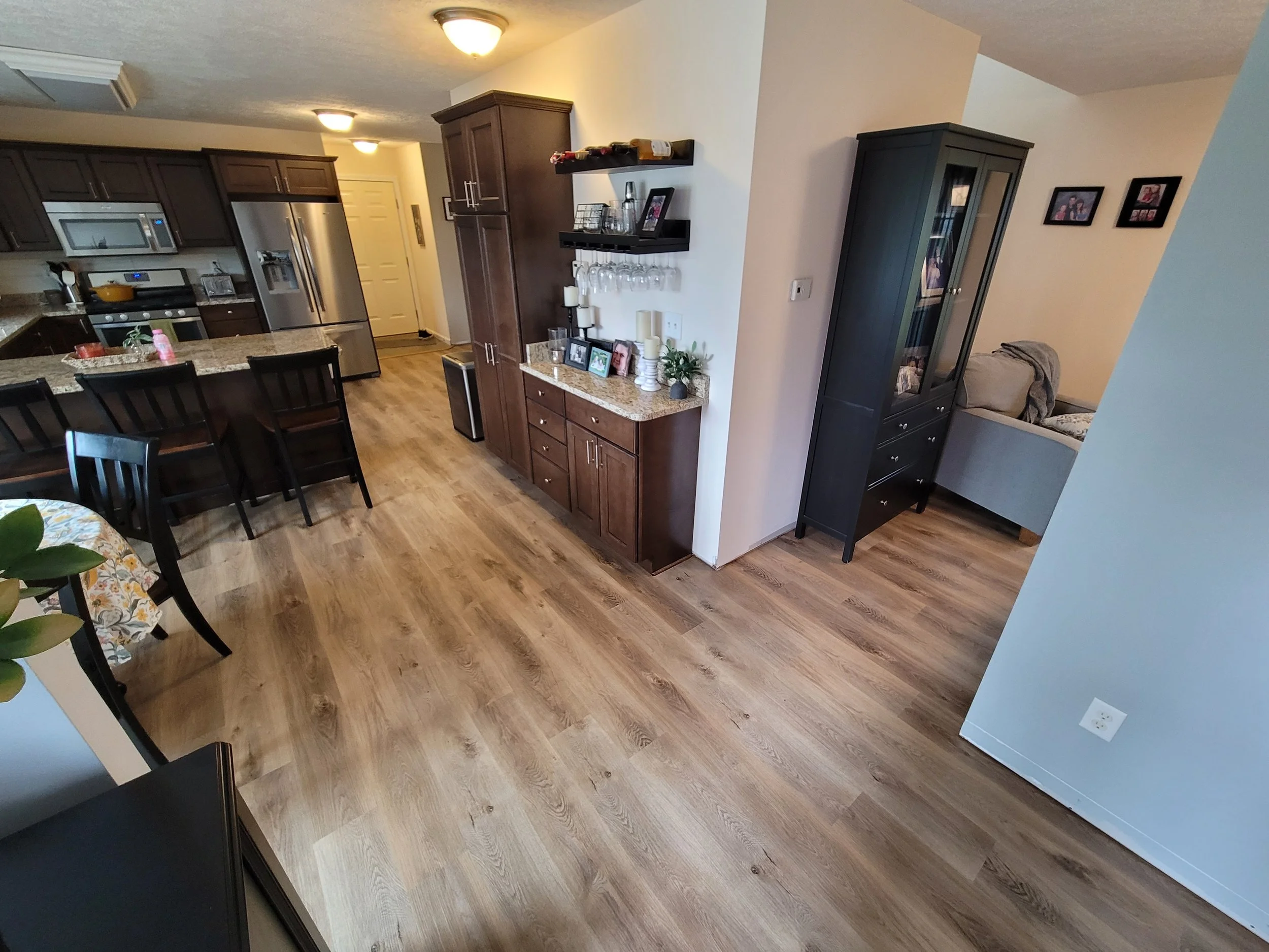 Interior view of a modern kitchen and dining area with wooden flooring, dark brown cabinets, a refrigerator, oven, and microwave, with a view into a living space with wall art and a gray sofa.