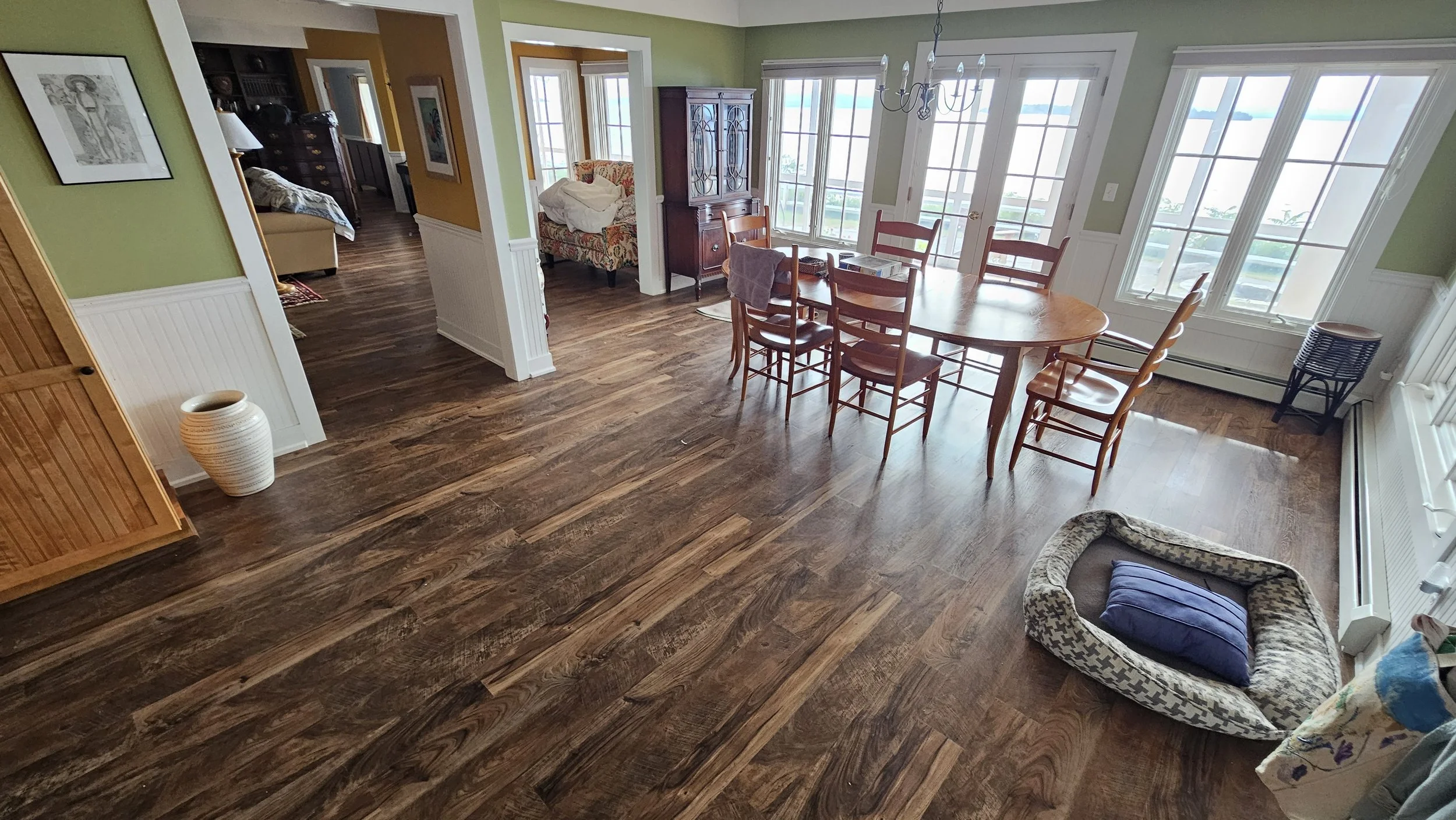 A dining area with a wooden dining table and six matching chairs. Large windows let in natural light. Other furniture includes a china cabinet, small side table, and a patterned pet bed on the floor. The flooring is hardwood.