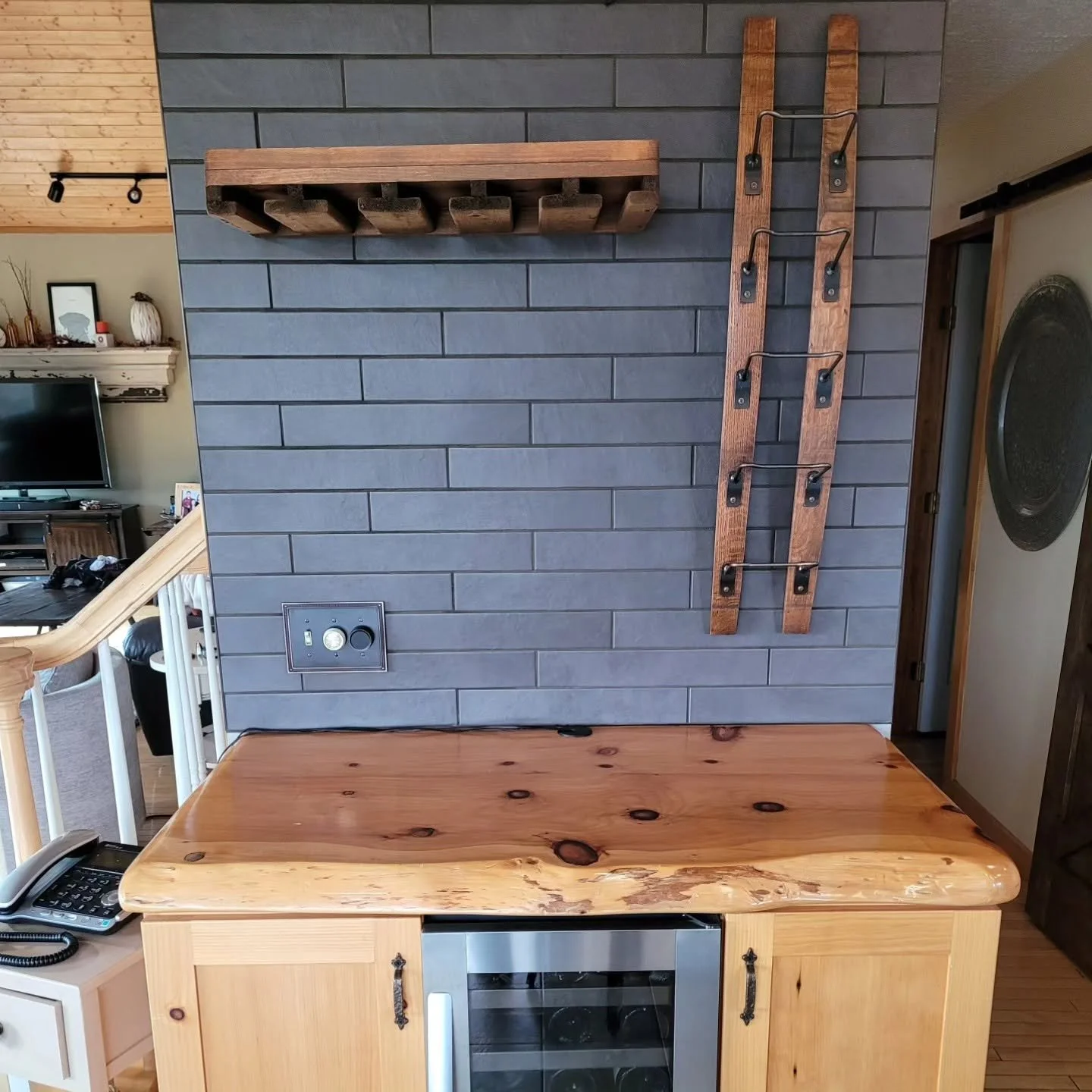 Interior of a home showing a gray brick wall with a wooden shelf, a vertical wooden ladder with metal hooks, and a built-in wine fridge below a wooden countertop. Part of a living area with a TV and decorations is visible in the background.