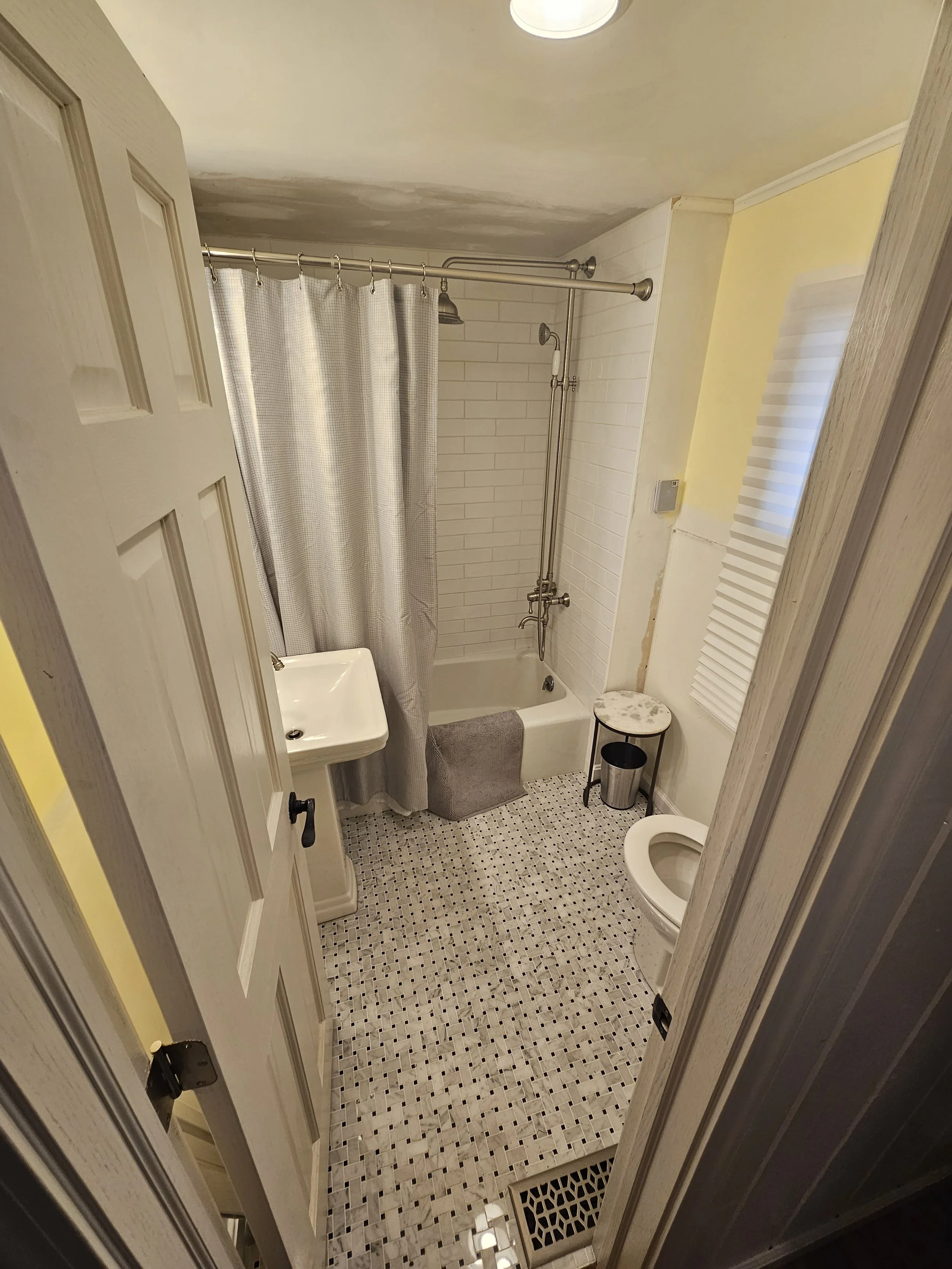 Small bathroom with a shower stall, white brick wall, beige ceiling, small square mosaic tile floor, pedestal sink, toilet, gray rug, small marble-topped table, metal trash can, window with blinds, and ceiling light.